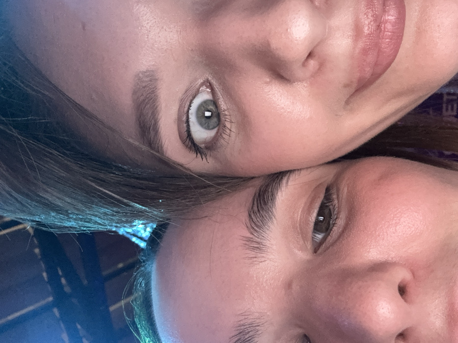 This image is a close-up portrait featuring two individuals.
**Subjects:**
1. **Left Individual:** Visible elements include the right side of a face, with fair skin, a discernible right eyebrow, a partially closed brown right eye, and the bridge of the nose. This individual's head is positioned to the left of the frame, lower than the second individual.
2. **Right Individual:** Visible elements include the right side of a face, with fair skin, a wide-open light grey-green right eye exhibiting a small, rectangular light reflection in the pupil, a defined right eyebrow, the right nostril, and a portion of the mouth revealing a slight smirk. Brown hair falls across the upper right portion of the frame. This individual's head occupies the right side of the frame, positioned higher than the first individual.
**Interaction:** The two individuals are in very close proximity, with their faces nearly touching, suggesting an intimate or playful interaction.
**Environment:** The background is dark and out of focus, showing blurred blue and black elements that suggest an indoor setting with artificial lighting, possibly at night. In the lower right corner, a purple fabric or object is partially visible, bearing white text that appears to spell "PRIVET" (Russian for "hello").
**Location:** The specific location within the city of Moscow, Russia, cannot be determined from the visual evidence provided.