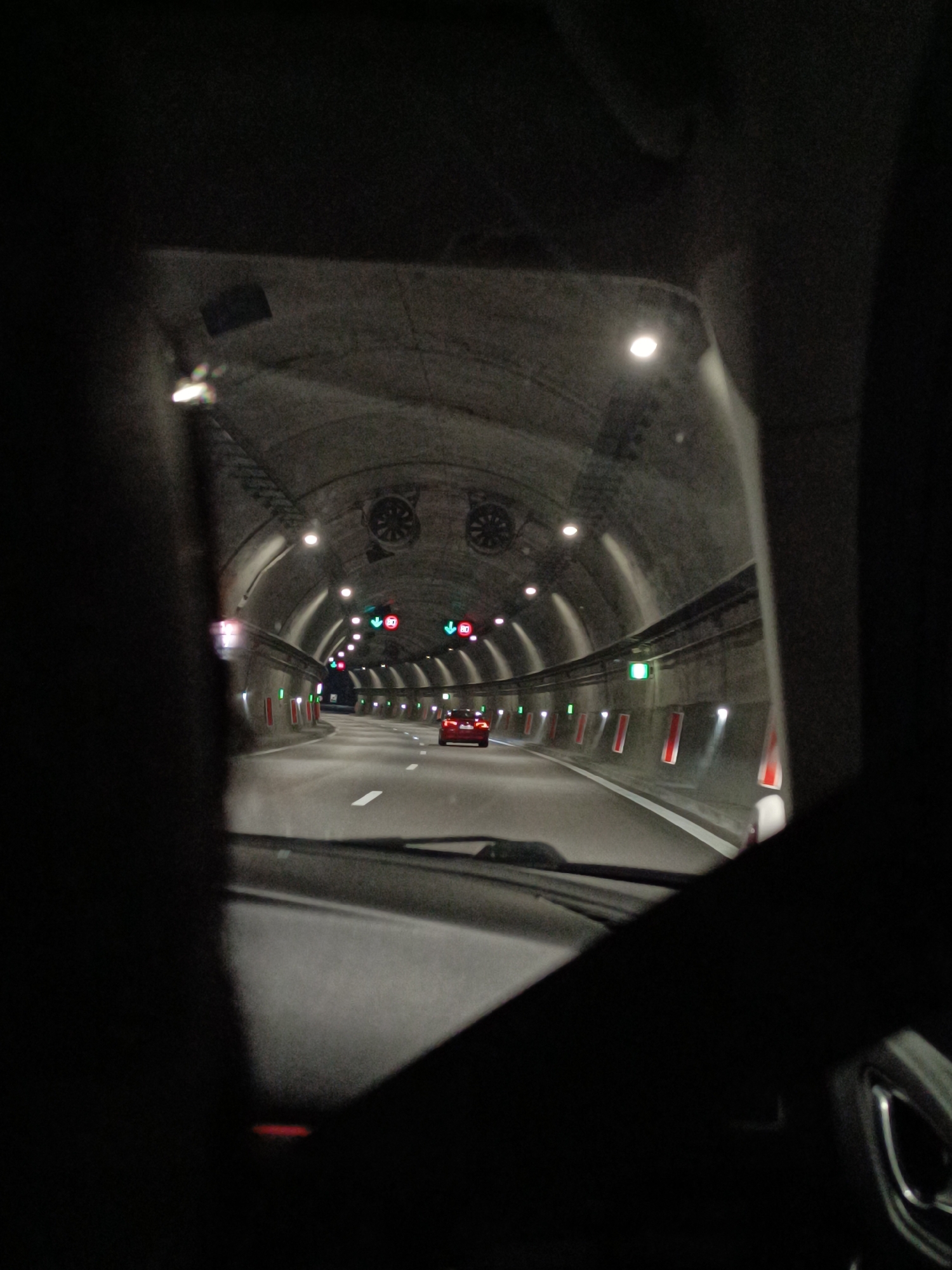 The image depicts a tunnel interior, possibly a highway tunnel, viewed from inside a vehicle. The tunnel has a gray concrete arched ceiling with regularly spaced circular lights and large ventilation fans. A red car is visible further down the tunnel, on a multi-lane road marked with white lines. The tunnel walls have green indicators and red reflectors. The foreground shows the interior of the vehicle, including the dashboard, windshield, and the steering wheel, all largely obscured by darkness. The location is unknown.