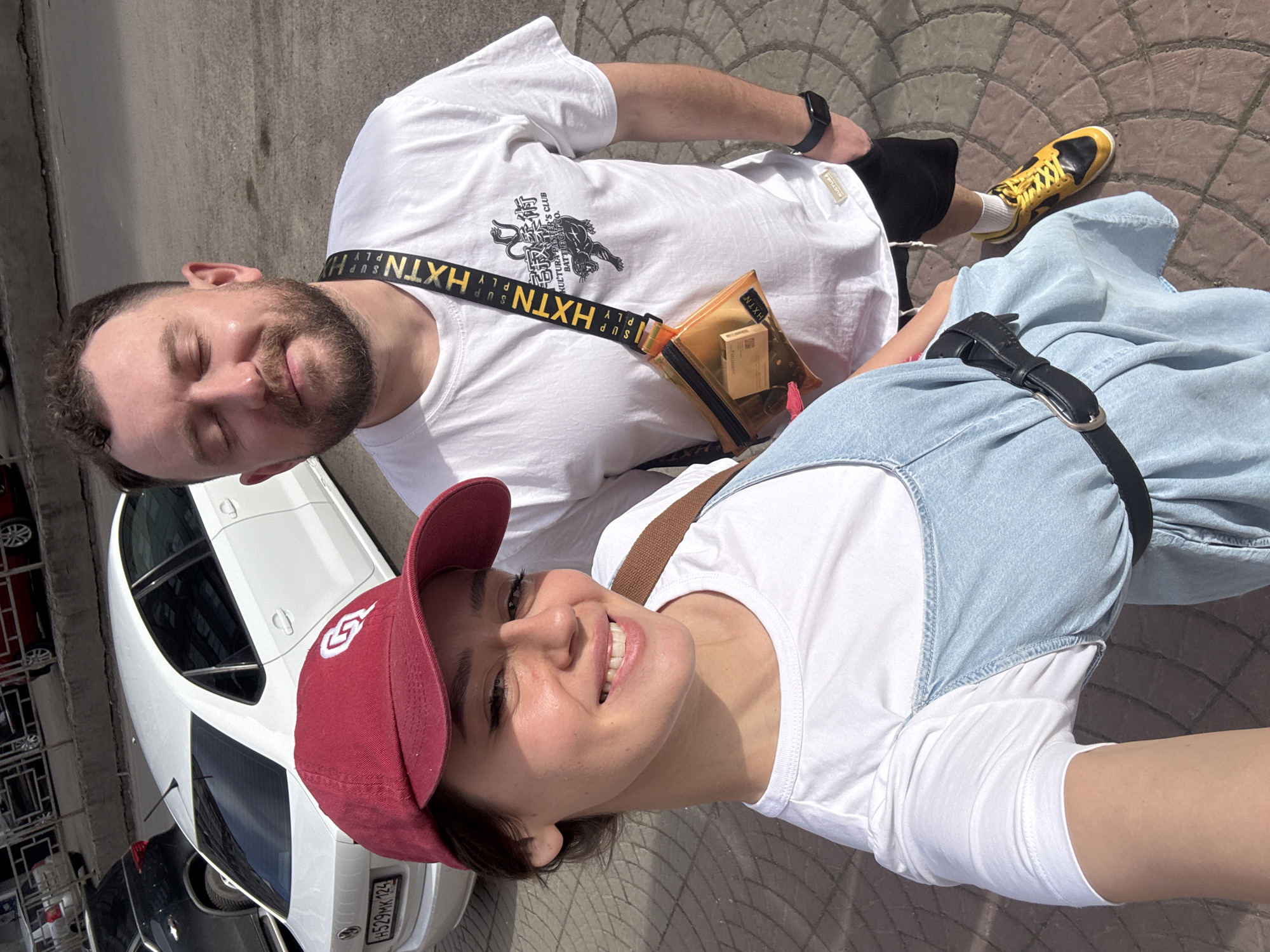 A selfie taken by a woman wearing a red baseball cap, a white t-shirt, and a light blue denim dress, capturing her and a man. The man, positioned to the right of the woman, is wearing a white t-shirt with a print featuring the words "Kultura Trader's Panter's Club" and the Japanese characters "宅寂柔術," black shorts, and yellow and black sneakers. He also has a black watch on his left wrist and a transparent orange crossbody bag with a black strap bearing the letters "HXTN" in yellow.
Behind them, a white car is partially visible, with its license plate reading "H529MK 124," indicating it is registered in Russia. The location appears to be outdoors on a paved area, likely a sidewalk or parking lot. Based on the car's license plate, the location is within Russia, specifically, Krasnoyarsk.