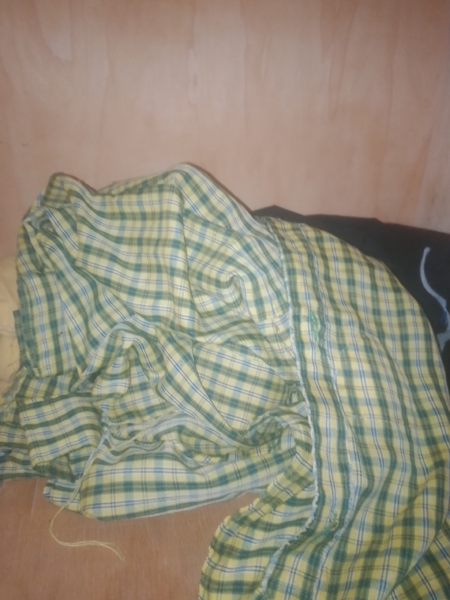 The image shows a wrinkled, light-colored plaid shirt with green and yellow checkered patterns, draped over a surface that appears to be a wooden floor or table. The shirt is not being worn. The background consists of a textured, light-brown wall. The scene is indoors, with no clear indication of its specific location within Jauro Yinu, Nigeria, other than the general domestic setting. The lighting is dim and appears to be artificial. No people are visible in the image.
