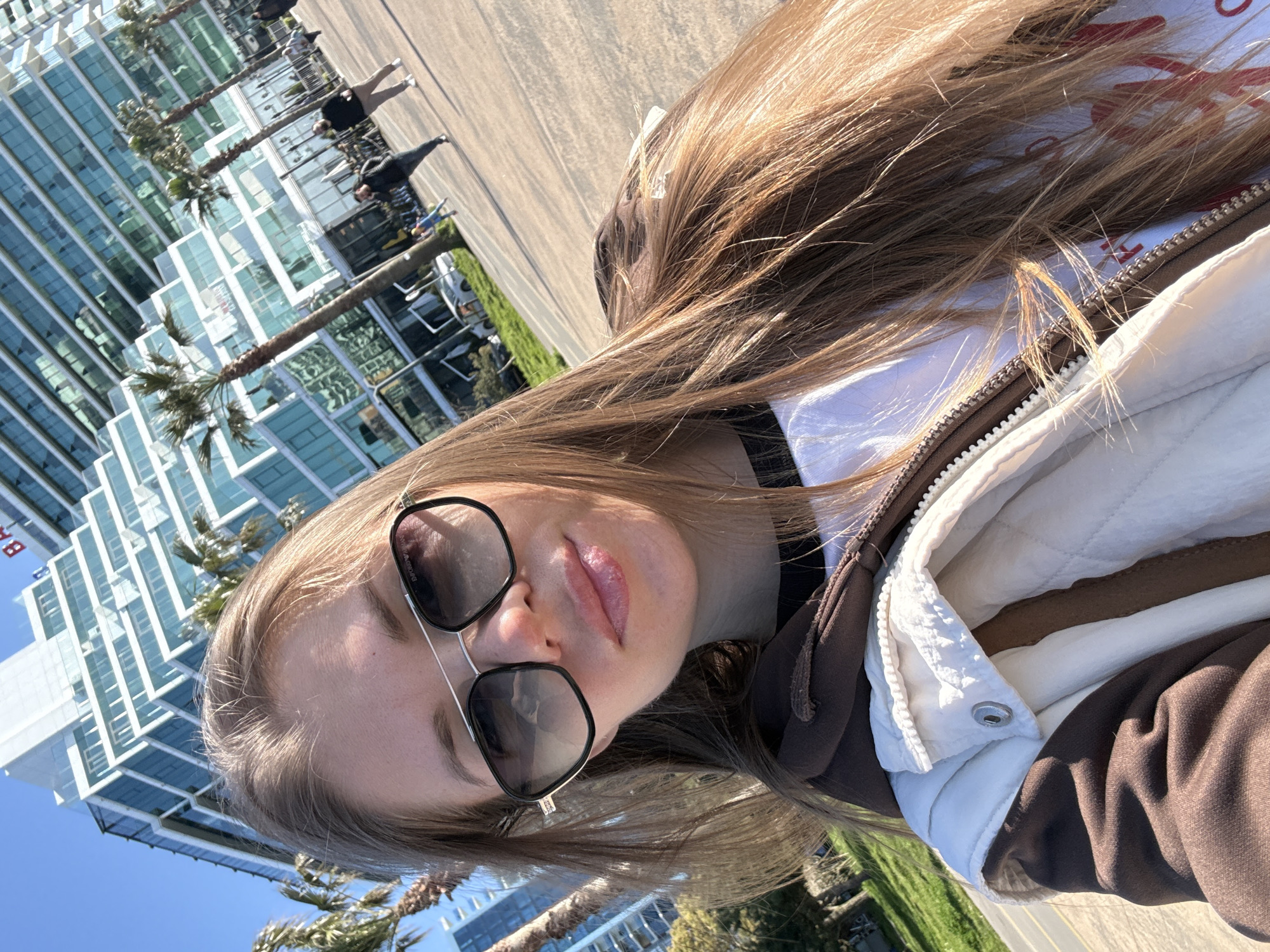 A young woman is captured in a selfie, positioned with her head slightly tilted and looking towards the camera. Her long, light brown hair, highlighted by the bright sunlight, cascades around her shoulders. She is wearing dark sunglasses that reflect the surroundings, and her lips appear to have a light gloss or lipstick. Her attire consists of a white top, a brown zipped hoodie or jacket, and an outer white quilted vest or jacket.
The scene is set outdoors on a very bright, sunny day with a clear blue sky. In the background, modern, tall glass buildings are prominent, reflecting the sky and surrounding structures. Several palm trees line the paved area, which could be a promenade or sidewalk. Faintly visible in the distance are a few other people; one distinctive figure appears to be a person in an animal costume, possibly an ostrich, and another person walking nearby, suggesting a public, tourist-friendly area. Green patches of grass are also visible.
The overall impression is one of a bright, urban, and possibly coastal location, consistent with Batumi, Georgia. The time of day is clearly daylight, indicated by the strong shadows and bright illumination. The woman appears calm and relaxed. The letters "BA" are faintly visible on the side of one of the buildings in the background.