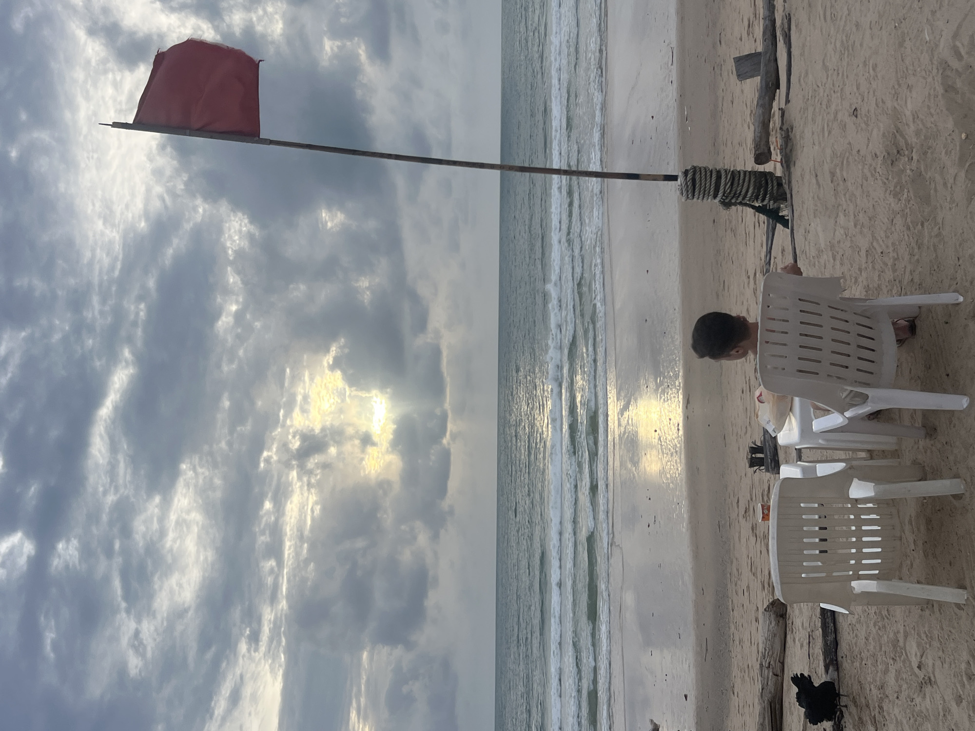 The image depicts a beach scene in Choeng Thale, Thailand, under a cloudy sky with sunlight breaking through. In the foreground, two white plastic chairs face the ocean. A small white table sits between them, with a few items on it, including what appears to be a white cup and a paper bag. A person, seen from behind, is seated in the chair on the right, facing the sea. To the left of the chairs, a black bird stands on the sand. Further back, a red flag on a wooden pole is planted in the sand. The beach is made of sand, with some driftwood scattered around. The ocean waves are visible, and the wet sand reflects the sky.