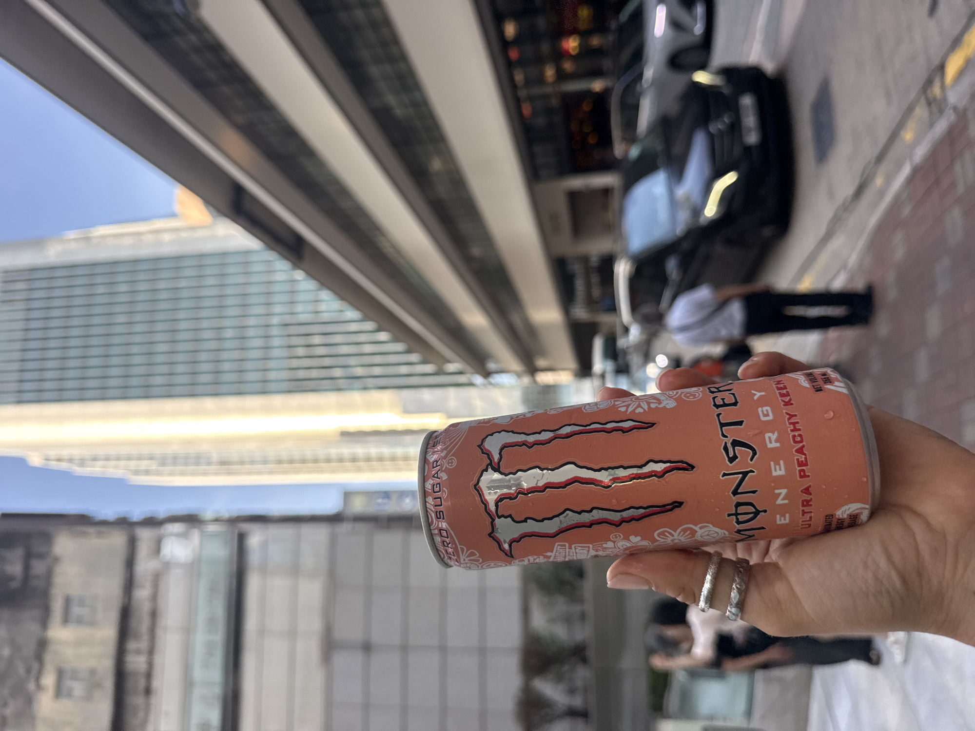 A close-up shot shows a hand holding a can of Monster Energy drink, "Ultra Peachy Keen" flavor. The can is orange with the Monster logo prominently displayed in silver and red. The background is blurred but suggests an urban environment with tall buildings and a street with cars. The hand has two rings on the ring finger and a tattoo on the wrist. The scene appears to be outdoors during the day, with the sky visible. Based on the architectural style of the buildings and the general ambiance, the location is likely Hong Kong, China.