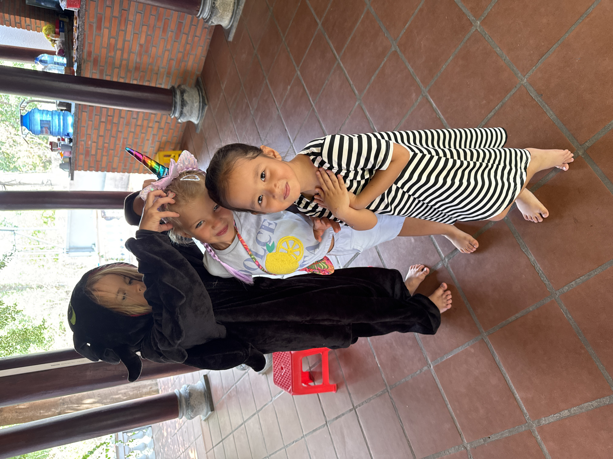 Two young girls are captured in a portrait, likely indoors or in a covered outdoor space in Mũi Né, Vietnam. The girl on the left, with blonde hair, wears a black, plush, full-body animal or character costume (possibly a dragon, given the green eyes on the hood) and a vibrant rainbow unicorn horn headband with a pink bow. Beneath her costume, a light-colored t-shirt with a yellow lemon graphic and partially visible text (likely "LEMON" or "LEMONADE") can be seen. She is touching her unicorn horn with one hand.
The girl on the right, with dark hair, wears a black and white striped dress or top. She has her hands clasped in front of her. Both girls are looking towards the camera with playful smiles. Their bare feet are visible on the reddish-brown tiled floor.
The background features brown architectural pillars on the left and in the mid-distance. Behind these pillars, a brick wall is visible with various items, including large blue water cooler bottles and smaller containers on shelves. A small, bright red plastic stool is positioned near the blonde girl's feet. The scene is well-lit, indicating it is daytime. The overall mood is cheerful and candid.