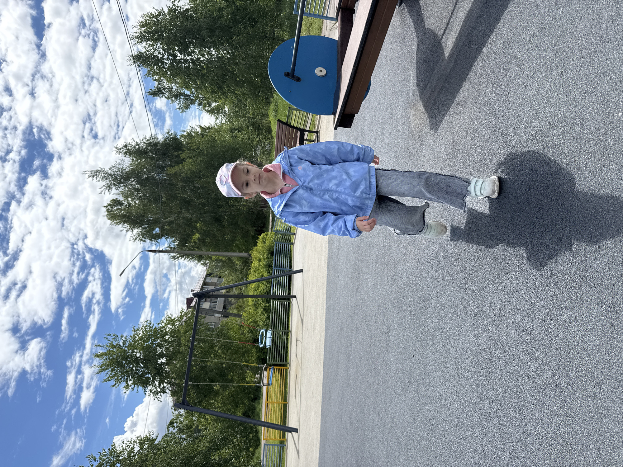 A young girl stands in a playground. She is wearing a blue jacket, gray pants, and a hat. Behind her is a playground structure, a swing set, and trees. The sky is blue with white clouds. The location is likely in Shevlyakovo, Russia, based on the prompt.