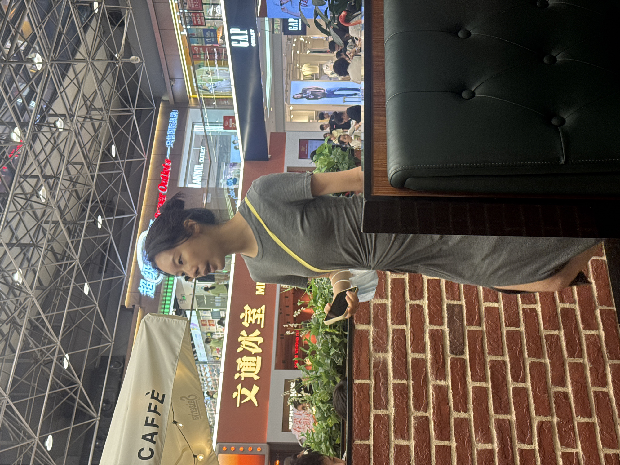 Here is a description of the image:
The image is taken at a shopping center or mall in Haizhu District, China. A woman wearing a gray dress and a yellow strap is looking at her phone. She is seated at a table, and there is a dark green cushioned bench behind her. A brick wall is adjacent to her. Outside, there is a store sign written in Chinese characters that says "文通冰室". Other store names visible are: CAFFÈ, ANNIIL OUTLET and GAP OUTLET. The roof of the mall has a crisscross metal structure.