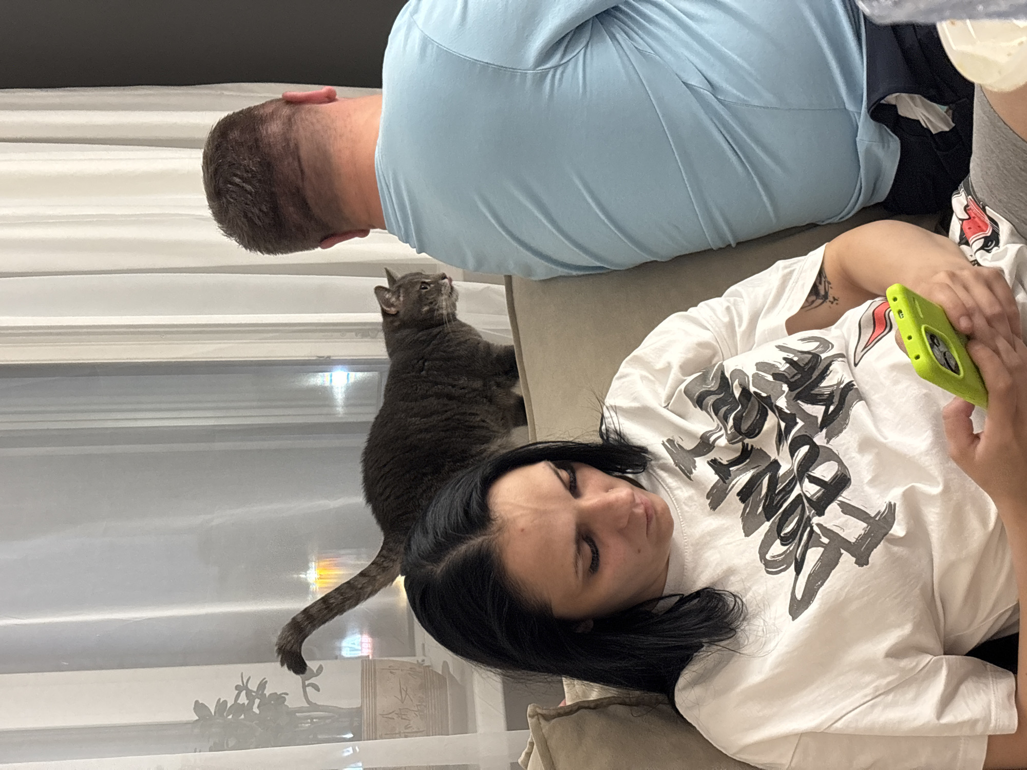 A woman with long black hair and dark eyes is looking down at her phone, holding it in her right hand. She is wearing a white t-shirt with a black graphic design. Beside her, a man with short brown hair is seen from the back, wearing a light blue t-shirt. He is sitting on a couch. Behind them, a tabby cat is walking on the back of the couch, with its tail raised. The scene appears to be indoors, with sheer curtains and a window visible in the background, suggesting it's nighttime or the blinds are drawn. The location is identified as Шебунино, Russia.