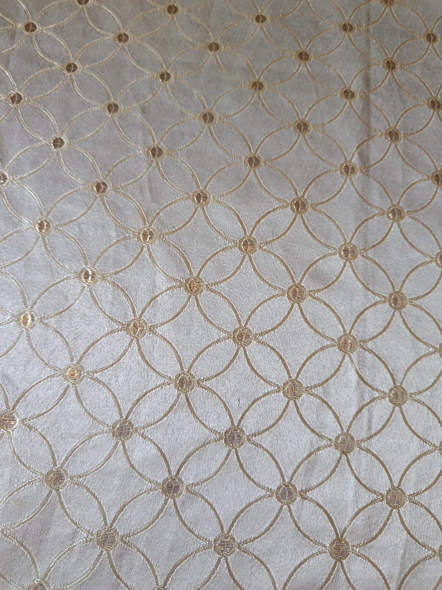 The image displays a close-up, overhead view of a textured fabric with a repeating pattern of interlinked circles. The fabric appears to be a light, neutral color, possibly cream or pale gold, and has a subtle sheen. The pattern is created by thin, embroidered lines in a slightly darker shade, forming a grid of overlapping oval shapes. Within each intersection of the ovals, there is a small, circular embellishment, also in a metallic thread. The overall impression is one of delicate ornamentation and refined texture. The image was taken in Гулбог 1, Tajikistan, and the specific location within the city is not discernible from the provided image.