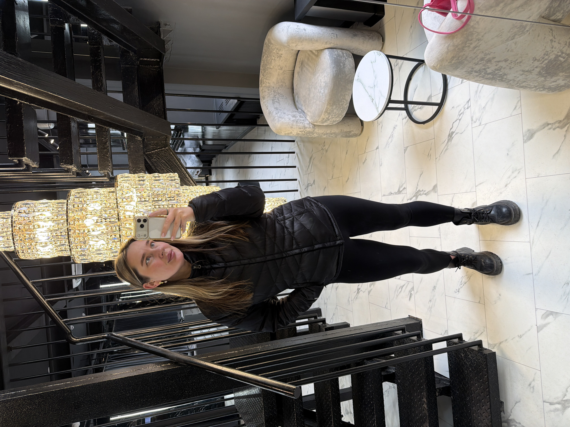 A woman is taking a selfie while standing on stairs with a black metal frame and railing. She is wearing a black quilted jacket, black leggings, and black boots. A large, multi-tiered crystal chandelier is visible above and to the left of her. Behind the woman is a staircase with a black metal frame. To the right, the floor is white marble, and there are two light gray chairs and a small marble-topped table. Based on the interior design, it could be located in Moscow, Russia.