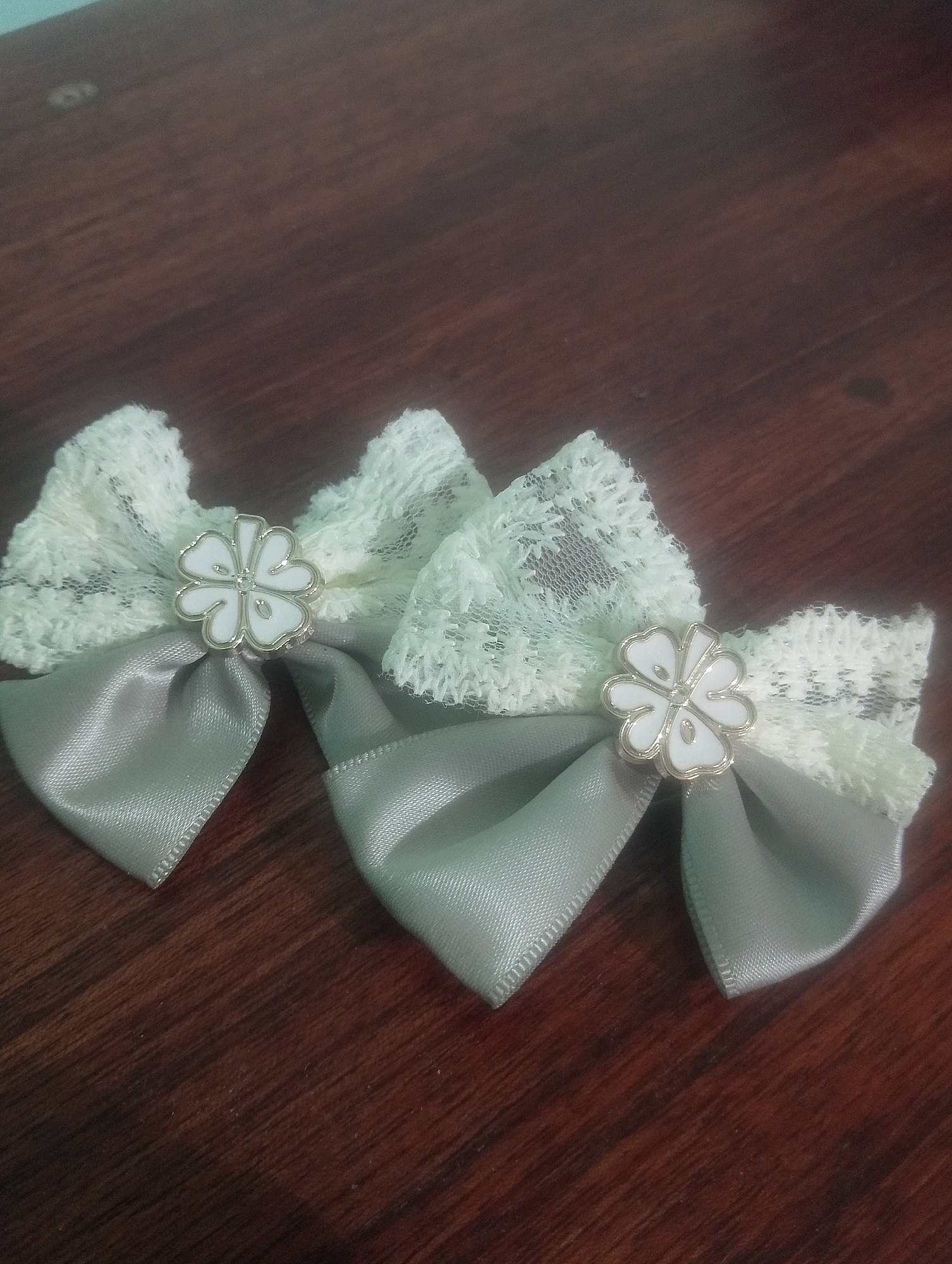 Two lace and satin hair bows with clover-shaped embellishments are displayed on a wooden surface. The bows are positioned in the foreground, with their tails extending downwards. The background is a dark, wooden tabletop with visible grain. There are no people or animals present. The scene appears to be indoors, possibly on a desk or table. The image does not provide any information that would suggest a location in Serbelawan, Indonesia, or any other specific city.