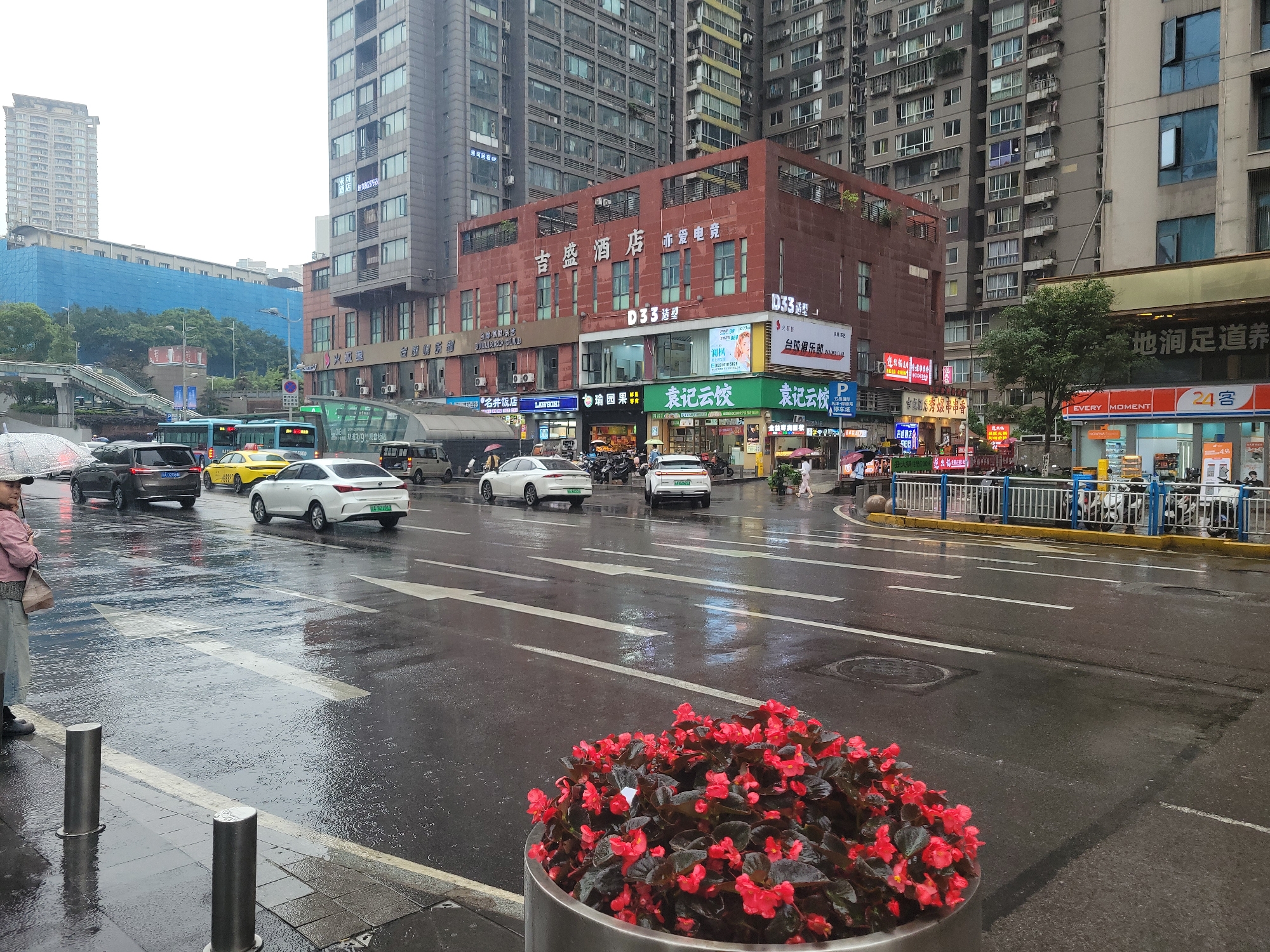 This image depicts a bustling urban street scene in Liangjiang, China, on an overcast and wet day. The asphalt road is slick with rain, reflecting the surrounding buildings and vehicle lights, with ripples visible on the surface from ongoing rain or recent showers.
The main subjects include numerous vehicles, such as cars (sedans, SUVs, electric vehicles), a yellow taxi, and a blue city bus, navigating the multi-lane street. Several pedestrians are visible on the sidewalks; one individual on the far left holds an umbrella, dressed in a light pink jacket and light-colored pants.
The setting is characterized by dense urban development, with numerous tall, modern high-rise buildings dominating the background, some appearing residential and others commercial. A prominent red-brown building in the mid-ground houses multiple businesses. Visible text on the storefronts includes "吉盛酒店" (Jisheng Hotel), "亦爱电竞" (also loves e-sports), "D33造型" (D33 Styling), "台球俱乐部" (Billiard Club), "名井饭店" (Mingjing Restaurant), "LAWSON" (convenience store), "白瑜园果" (Baiyu Garden Fruit), and "袁记云饺" (Yuanji Yunjiao). Other signs advertise "地涧足道养" (Dijian Foot Spa & Health), "EVERY MOMENT," "24客," and "轨道3/9观音桥站" (Line 3/9 Guanyinqiao Station), indicating proximity to a subway/monorail station in Chongqing. License plates, such as "A 824913," are also visible, consistent with Chongqing.
On the left, a large building is shrouded in blue construction netting, with a pedestrian overpass visible beneath it. In the foreground, a planter bursts with vibrant red flowers, adding a splash of color to the otherwise muted palette of the rainy city. The time of day is likely daytime due to the ambient light, though the overcast sky suggests poor visibility conditions. The scene captures the routine activity of an active city, with traffic and pedestrians moving despite the wet weather.