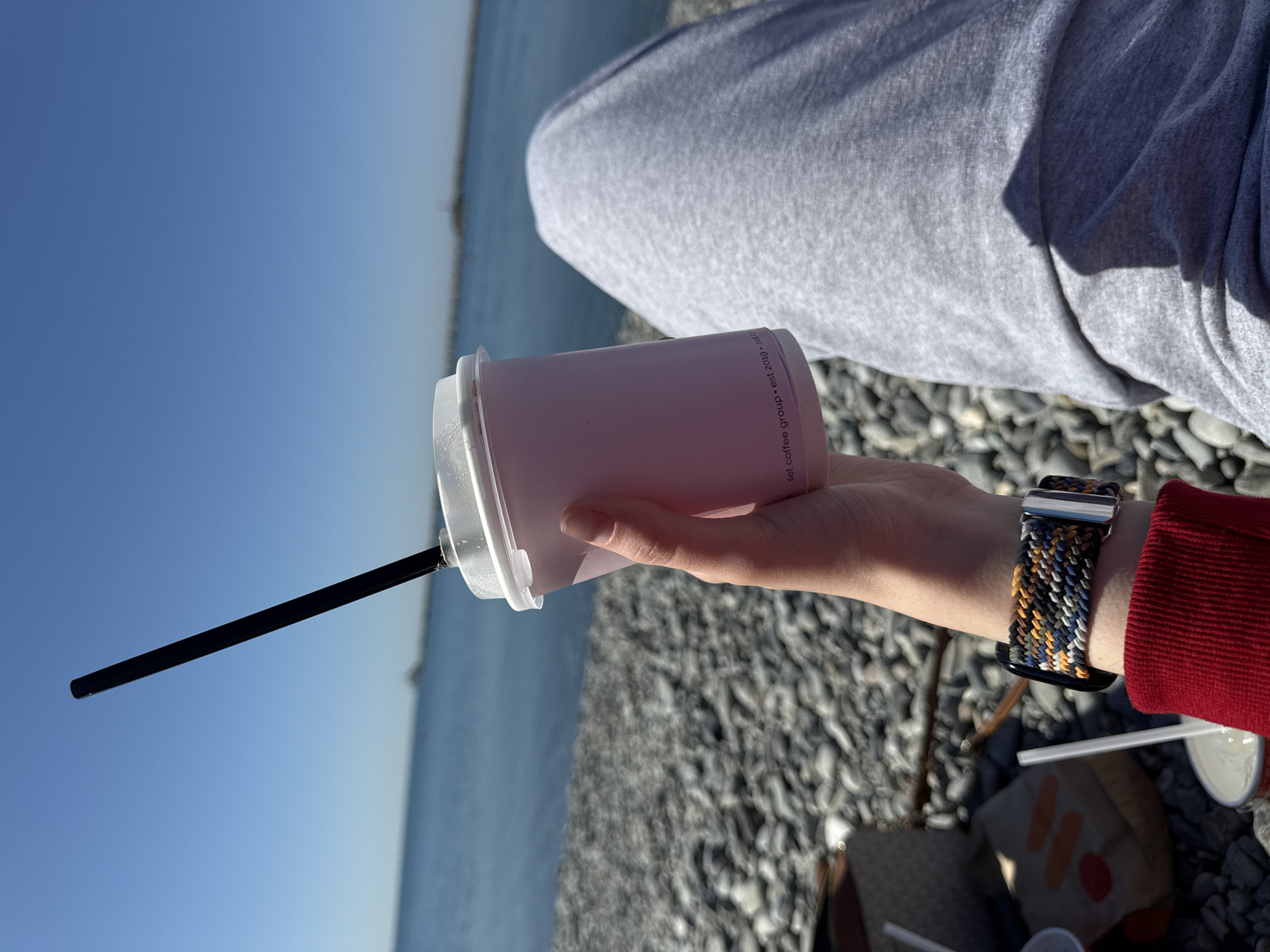 The image captures a person's perspective as they sit on a pebble beach, holding a drink. The main subjects are the person's hand, arm, and a leg clad in grey sweatpants, holding a light pink disposable cup with a clear lid and a black straw. A multi-colored braided watch band, featuring shades of blue, yellow, white, and black, is prominently displayed on the person's wrist, which is covered by a red sleeve.
The setting is a sunny outdoor location, with a vast, calm body of water (likely a sea or ocean) stretching to the horizon under a clear blue sky. The foreground and mid-ground are dominated by a pebble beach. The bright sunlight casts distinct shadows on the grey sweatpants and the pebbled ground, indicating a clear and sunny day.
The activity taking place appears to be a relaxing moment by the water, enjoying a beverage. Visible text on the pink cup reads: "sbl coffee group • est. 2019 •". Other blurry objects, possibly another cup or a bag, are visible on the ground beside the person.