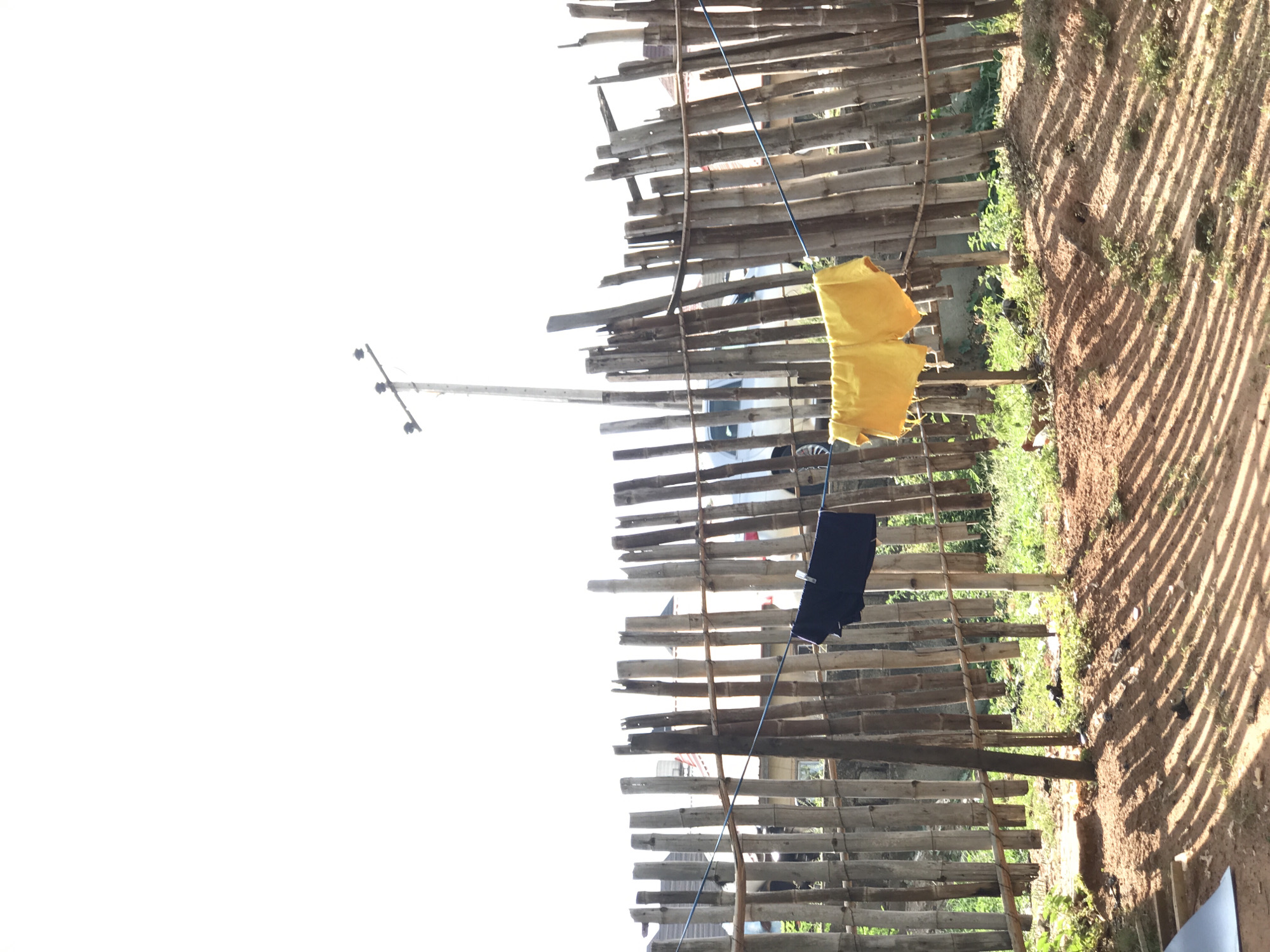 The scene depicts a bamboo fence, oriented vertically, with yellow and black clothes hanging on a clothesline. A utility pole is visible in the background. The ground beside the fence has dirt and sparse vegetation. The image was taken in Abuja, Nigeria.