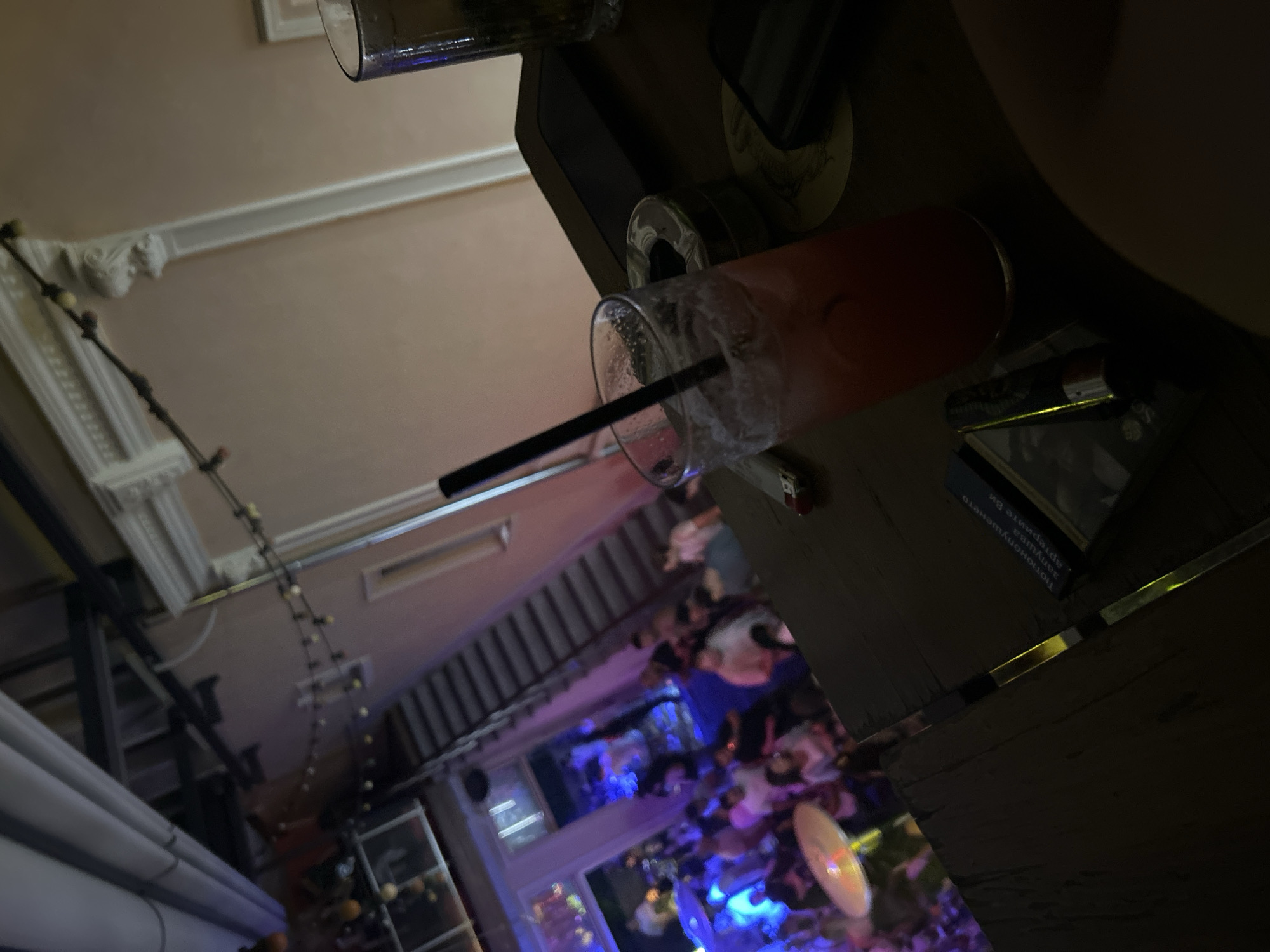 The scene is indoors at a bar or restaurant in Plovdiv, Bulgaria. A cocktail with a straw is placed on a dark-colored table, and in the background, there are many people illuminated with a blue and purple light effect. Decorations are on the wall on the left-hand side of the image.