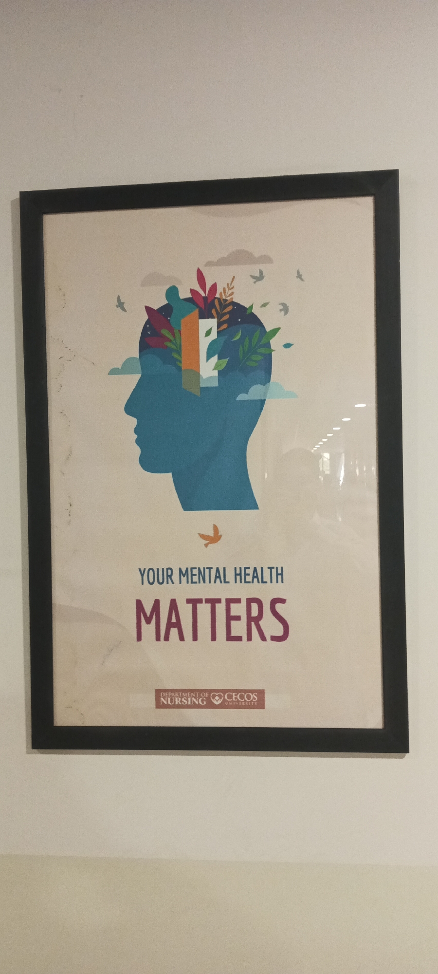 The image shows a framed poster hanging on a light-colored wall. The poster depicts a blue silhouette of a human head. Inside the head, there are illustrations of leaves, plants, clouds, and birds. Above the head, the text "YOUR MENTAL HEALTH" is printed in a light gray, and below that, "MATTERS" is printed in a dark purple-red color. At the bottom of the poster, there's a logo that reads "DEPARTMENT OF NURSING" with a heart symbol, followed by "CECOS UNIVERSITY". The location is inferred from the CECOS University logo, which suggests it's in Peshawar City Tehsil, Pakistan.
