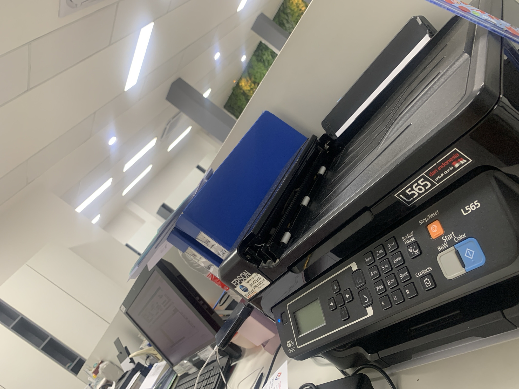 The image shows a close-up of a black Epson L565 printer on a desk. The printer has a control panel with buttons and a small LCD screen. There is also a sticker on the printer that says "EPSON" with a phone number. In the background, there is a computer monitor, a blue binder, and some shelves. The lighting suggests it is an indoor environment, possibly an office. The printer is labeled "L565" and "dari Indonesia untuk dunia," indicating its manufacturing origin. The image is taken at an angle, tilting the view of the printer and its surroundings.
