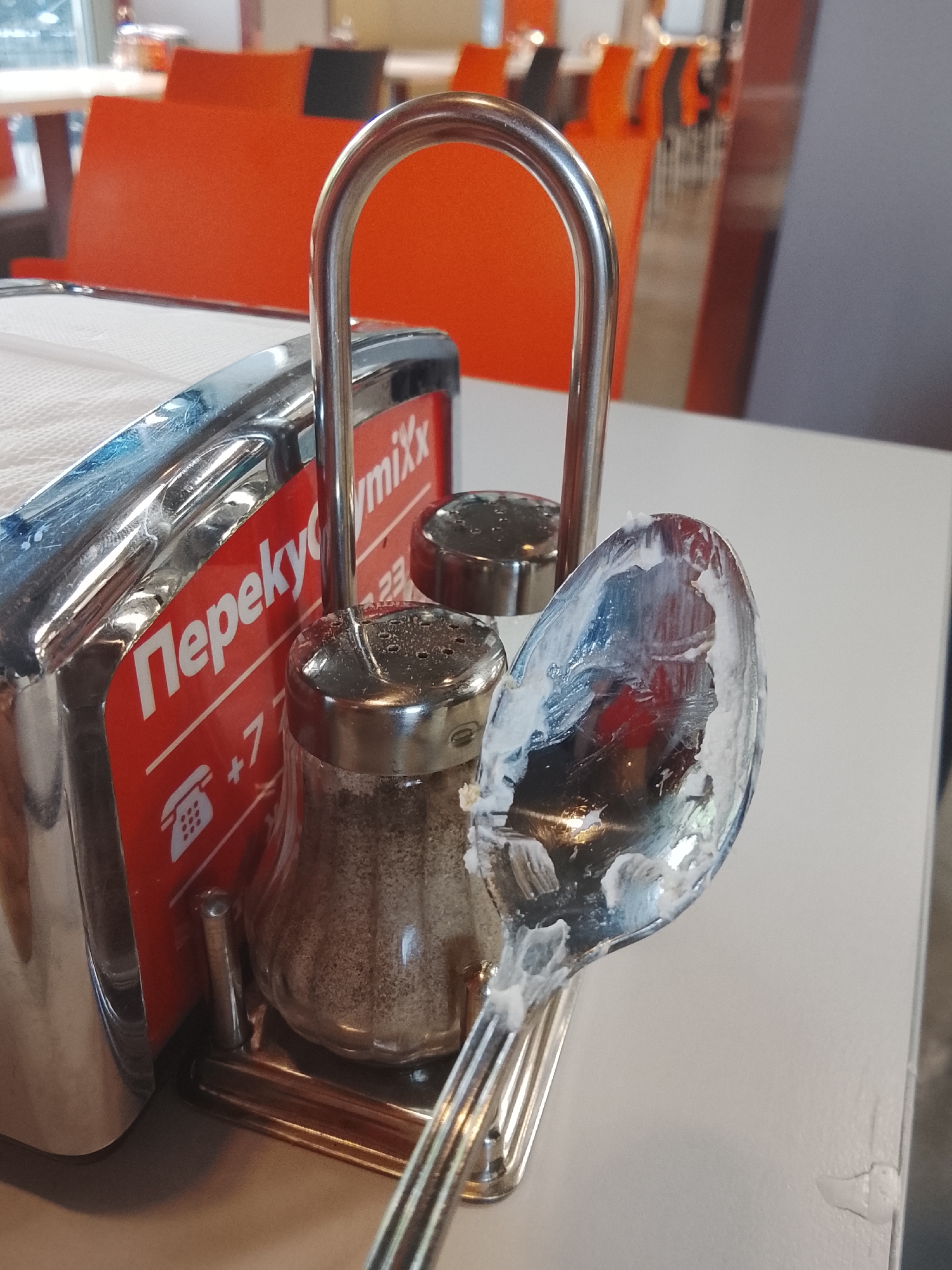 The image depicts a close-up of a table setting in what appears to be a casual dining establishment, possibly a cafe or fast-food restaurant. The scene is dominated by a metal napkin dispenser with a red label featuring white text. The text on the label is in Cyrillic and appears to be a brand name, "Перекууу" followed by "mixx" and some numbers, including a phone number starting with "+7". Next to the napkin dispenser are two salt and pepper shakers, one of which is partially filled with a dark granular substance, likely pepper. A shiny metal spoon, visibly coated with a white creamy substance, rests precariously on the edge of the table, positioned to the right of the shakers. The background suggests an indoor environment with orange seating and a hint of natural light coming from windows. While the image quality and angle do not provide definitive clues about the specific location within Almaty, Kazakhstan, the presence of Cyrillic text on the napkin dispenser is consistent with the region. The overall impression is one of a recently used dining setting, with remnants of a meal or dessert on the spoon.