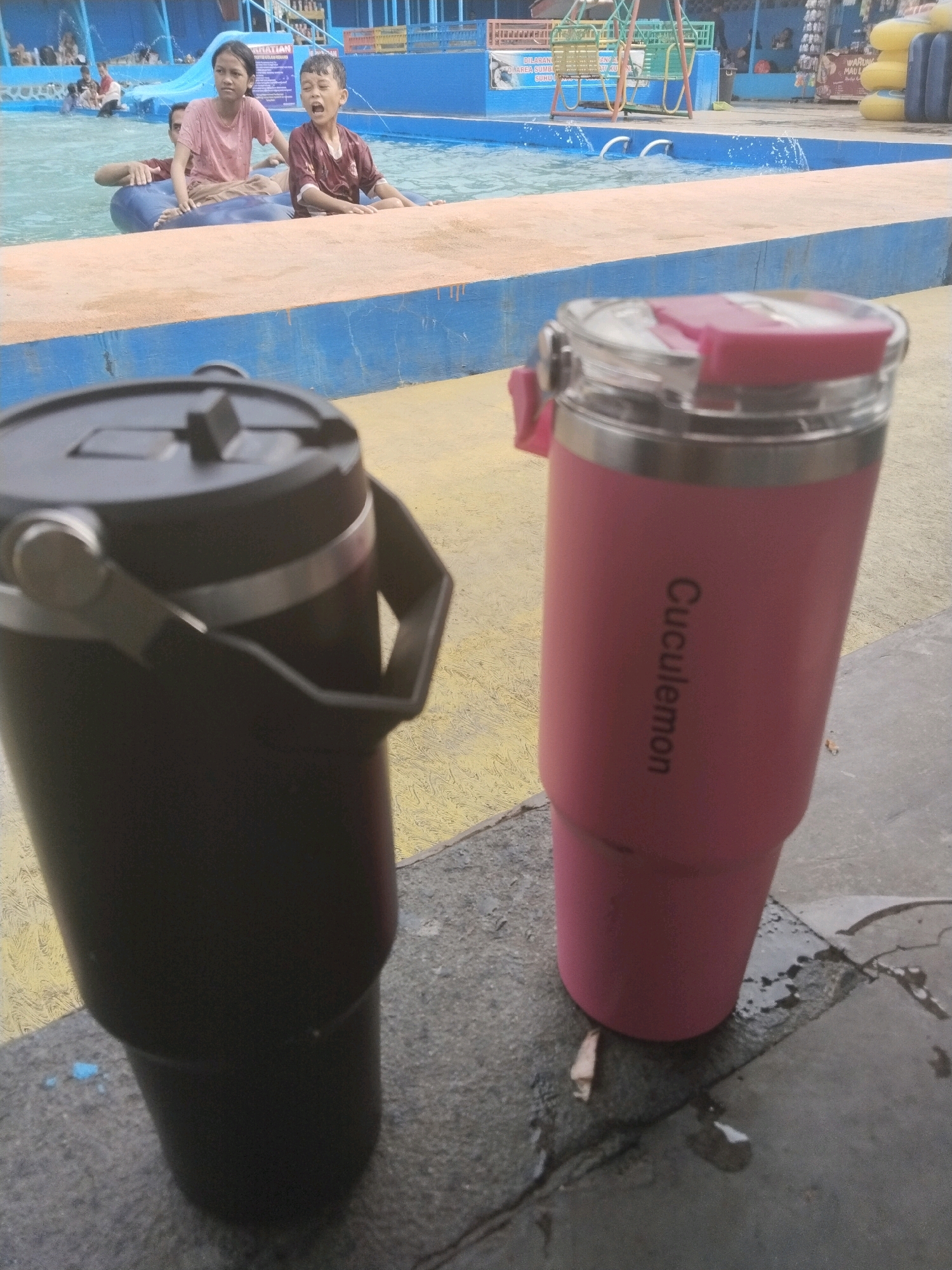 Two large beverage tumblers, one black and one pink, are in the foreground, resting on a wet concrete surface near the edge of a swimming pool. The pink tumbler has the word "Cuculemon" printed vertically on it. In the background, partially submerged in the blue water of the pool, are three people: a young boy in a maroon shirt with his mouth open, a young girl in a pink shirt, and a person behind them wearing a maroon shirt. The pool area appears to be part of a public facility, with a yellow and blue painted ledge separating the pool from the walkway. Further in the background, there are signs, a swing set, and other amenities typical of a water park. The scene is likely located at the Batukuwung Water Park in Indonesia, given the context of similar images.