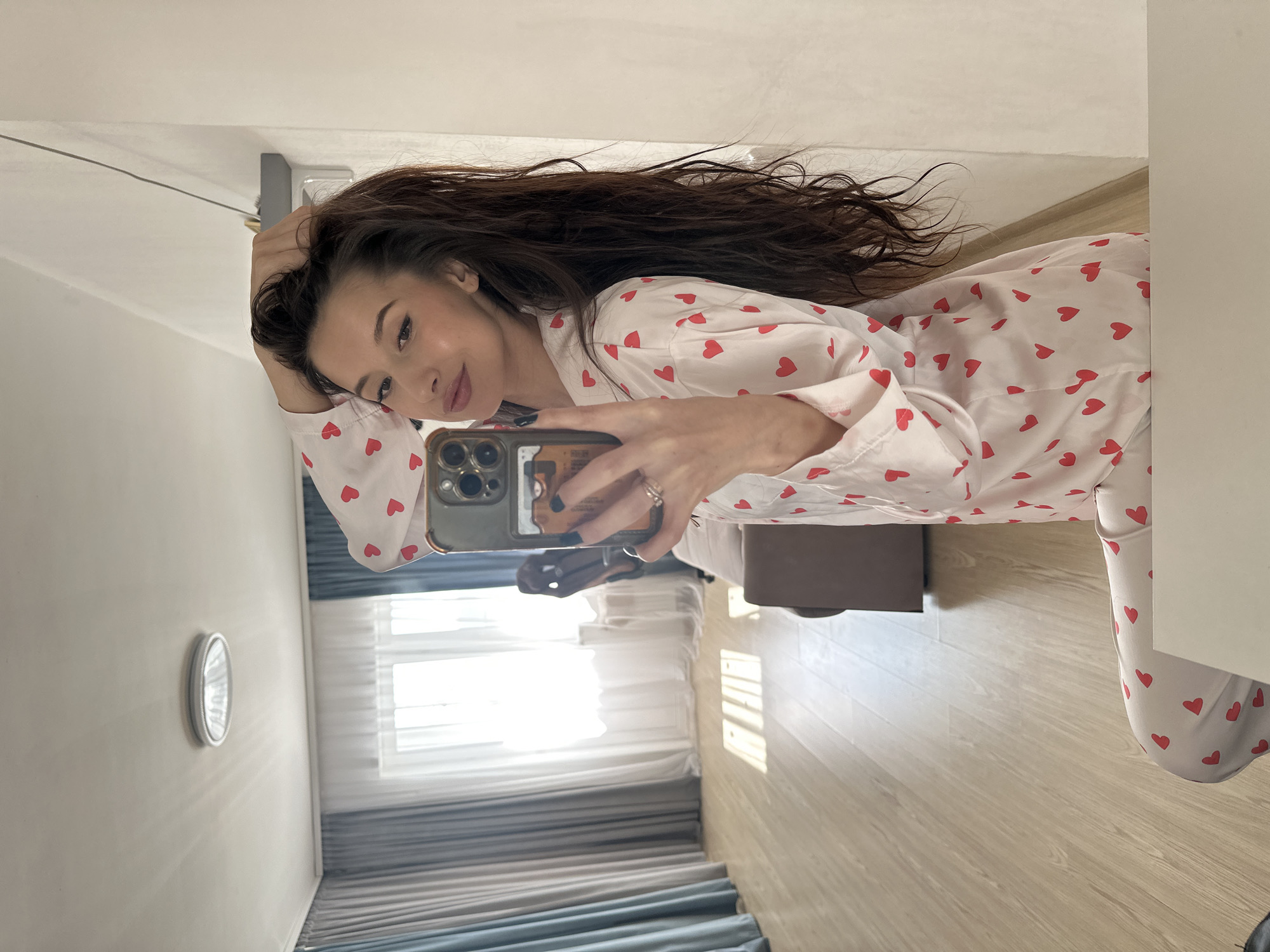 A woman in pajamas with red hearts is taking a selfie indoors. She is holding a smartphone in her right hand. She has long, dark, wavy hair. The background includes a room with curtains, a wooden floor, a cabinet, and a wall. It is not possible to confirm the location in Yekaterinburg, Russia based on the provided image.