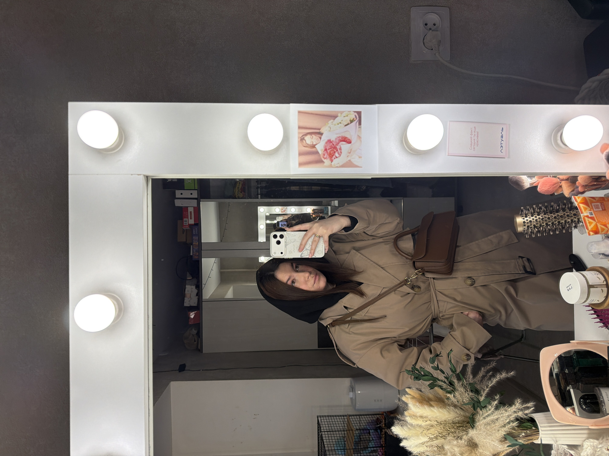 A woman is taking a selfie in a mirror surrounded by vanity lights. A light-colored trench coat and a small brown purse are visible. The woman has dark hair and is wearing a dark scarf or hood. Behind her, the reflection shows a room with shelves and various objects. The mirror has several items attached to the top, including a photograph and a small card. The location is likely an indoor setting, such as a dressing room. The city and country information cannot be verified by any objects in the image.