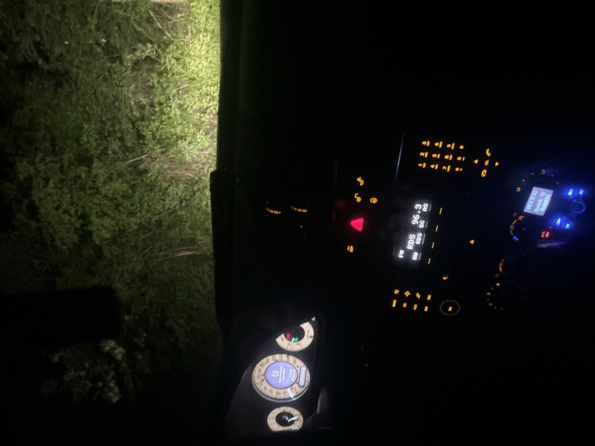 The image appears to be taken from inside a car at night, focused on the dashboard and a partial view outside the driver's side window. The dashboard is illuminated with various lights, including the instrument cluster, radio display, and warning lights. The radio display indicates the frequency 96.3 RDS. The instrument cluster shows the car's gauges and information panel. Outside the window, there is vegetation, possibly bushes or trees, illuminated by what seems to be headlights. The environment is very dark. No people are visible. There is no information from the image indicating that this image was taken in Veliko Tarnovo, Bulgaria.