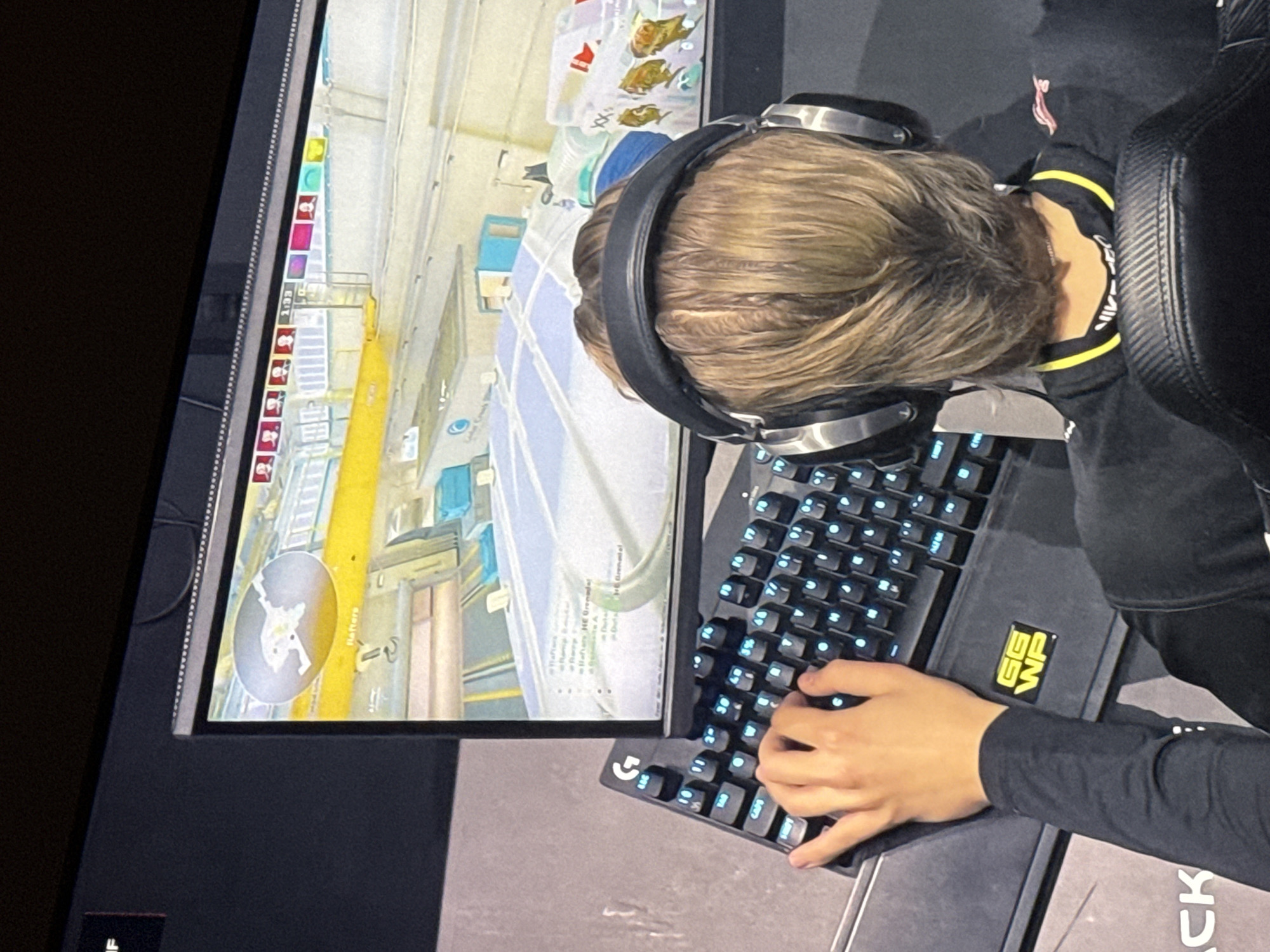 An overhead, medium shot shows a person wearing a headset and black long-sleeve shirt playing a first-person shooter video game on a computer. The game screen displays a map and player positions. The person's hands are positioned on a black keyboard with blue backlighting. A portion of a gaming chair is visible on the right side of the frame. No further location details are discernible from the image.