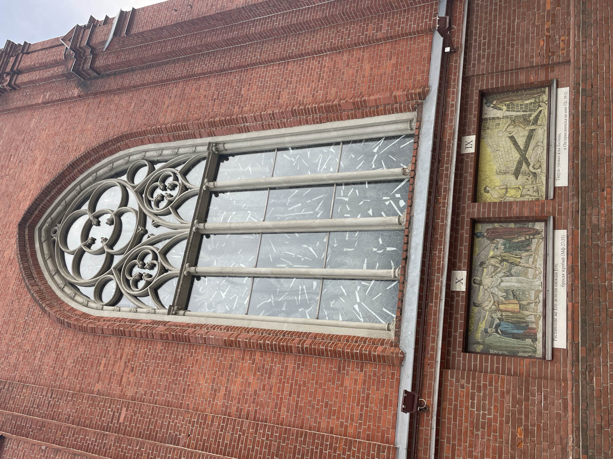 The image shows the exterior of a brick building, likely a church, with a large Gothic-style window dominating the upper portion. The window features intricate tracery and stained glass. Below the window, there are two horizontal mosaic panels. The left mosaic depicts a scene with several figures, and below it, a Russian inscription. The right mosaic shows a scene with a cross and figures, also with a Russian inscription below. The architecture suggests a European style, and the inscriptions are in Russian. The building is made of red brick and has a somewhat overcast sky visible in the background. The image was captured from a low angle, looking upwards at the building. Based on the Russian text, the building is likely located in Moscow, Russia. The mosaic on the left appears to depict Jesus being stripped of his garments, with the inscription "They divided His garments among themselves, casting lots" (Matthew 27:35). The mosaic on the right shows Jesus falling with the cross, with the inscription "I was firmly resolved to serve the Lord, and He inclined to me" (Psalm 39:1). These are likely scenes from the Stations of the Cross.