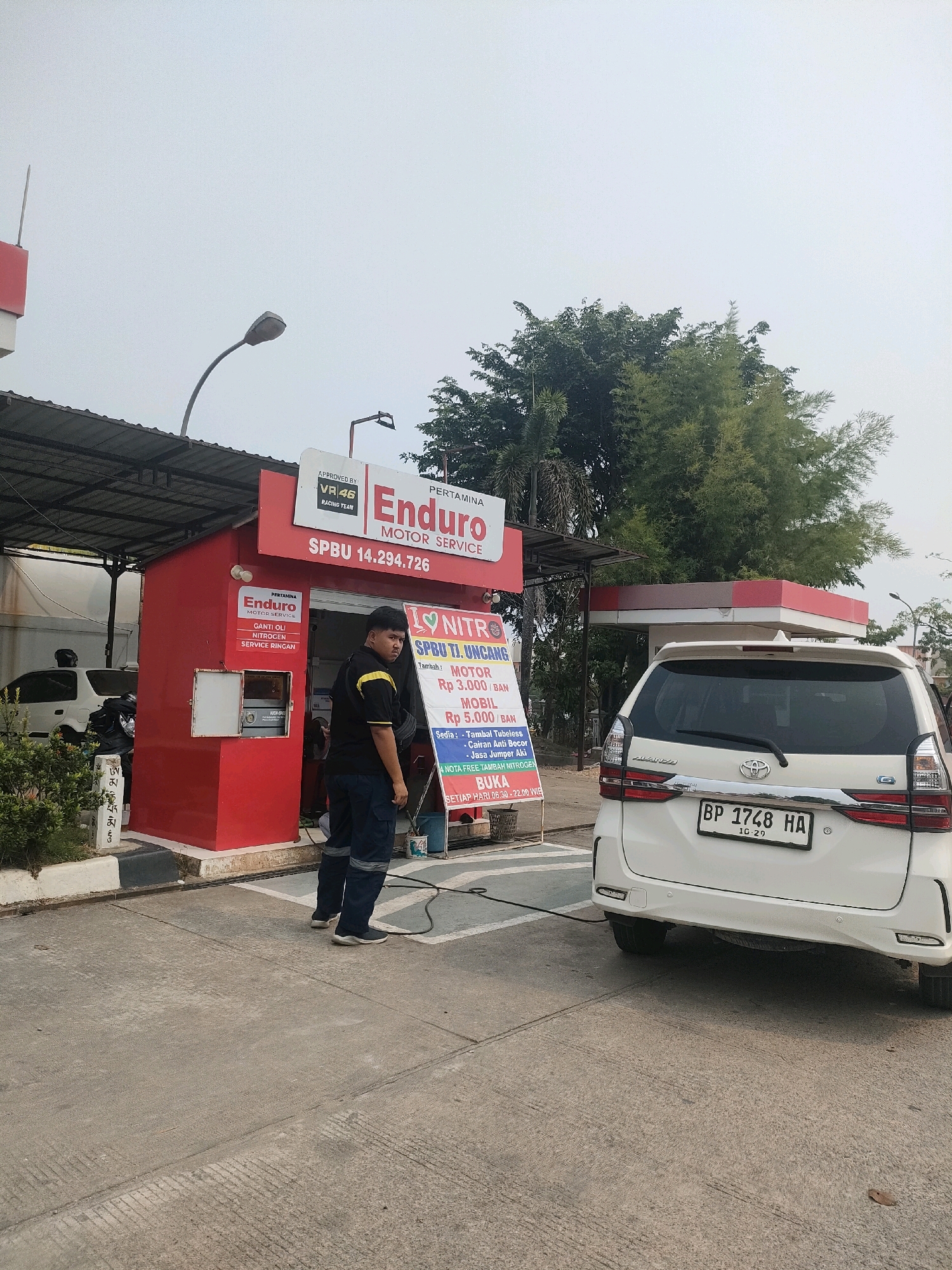 An outdoor scene shows a white Toyota Avanza with license plate BP 1748 HA parked at a Pertamina Enduro Motor Service station in Batam, Indonesia. A fuel hose from the station is connected to the rear of the car. Two men in work uniforms are present. One man, wearing a black shirt with yellow accents and dark pants, stands near the service station booth. The other man is partially visible behind the car. The service station is red with prominent signage for "Pertamina Enduro Motor Service" and "SPBU 14.294.726". A price list for services like "GANTI OLI" (oil change) and "NITROGEN" is displayed on the booth. Trees and an overcast sky form the background.