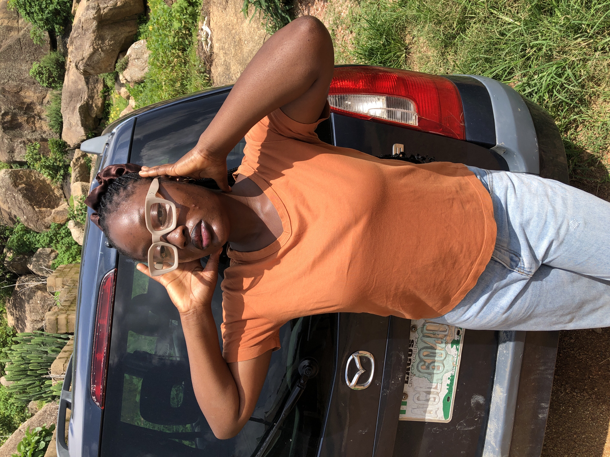 A young Black woman with dark skin and braided hair tied with a brown scrunchie poses in front of a dark blue SUV. She is wearing large, light-colored, square-framed glasses, an orange short-sleeved t-shirt, and light blue denim jeans. Her hands are raised to her head, with her fingers tucked behind her ears. The SUV has a silver Mazda logo on the back and a Nigerian license plate that reads "LAGOS CENTRE OF EXCELLENCE AGL 604D FEDERAL REPUBLIC OF NIGERIA". The background features rocky terrain with sparse green vegetation, including tall, succulent-like plants, and what appears to be the city of Jos, Nigeria. The lighting suggests it is daytime.
