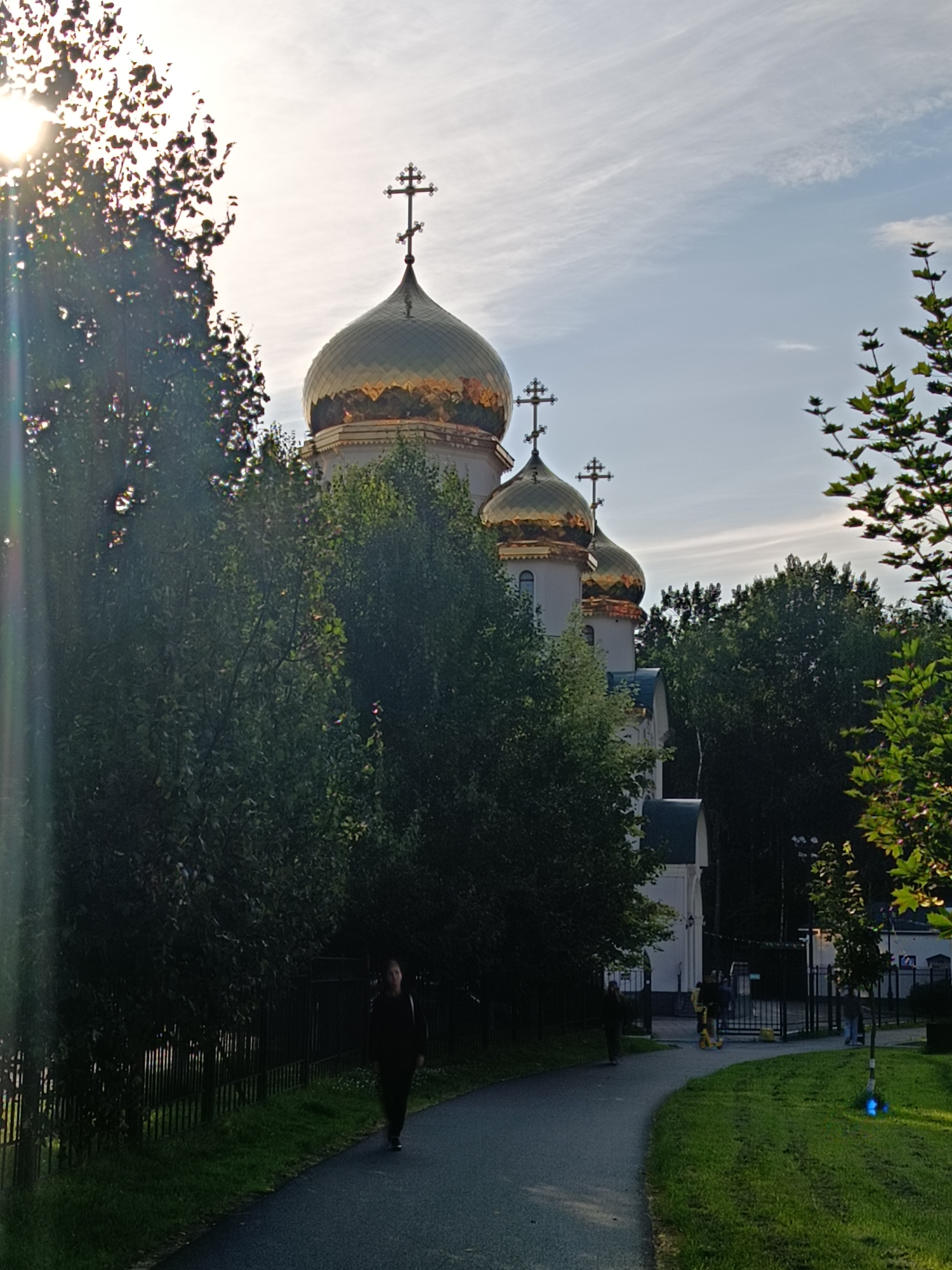 The image shows the golden domes of an Orthodox church, likely in Saint Petersburg, Russia, bathed in the warm light of the setting sun. The sun's rays are filtering through the branches of lush green trees, creating lens flare effects on the left side of the frame. In the foreground, a paved path curves through a grassy area. A lone figure, dressed in dark clothing, walks away from the camera on the path. Further along the path, near an entrance gate, several other figures are visible, including one on a yellow scooter. The church itself is partially obscured by trees, but its prominent golden domes and crosses are visible against a pale blue sky with wispy clouds. A dark metal fence runs along the edge of the path.