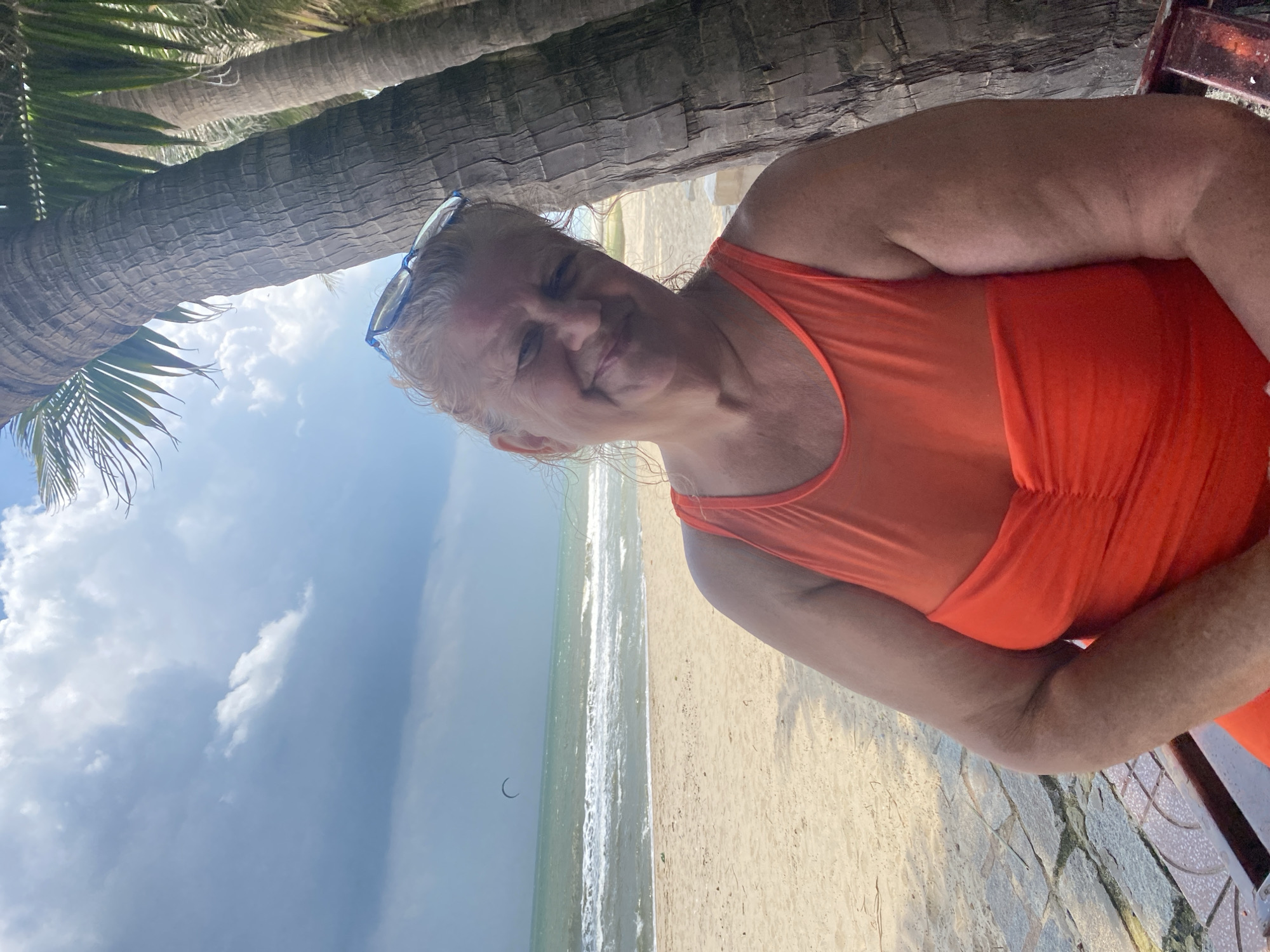 An outdoor, medium shot shows a woman sitting on a bench under palm trees on a beach in Mũi Né, Vietnam. The woman, who appears to be in her late 50s or early 60s, has blonde hair and is wearing an orange one-piece swimsuit with a sheer neckline. She is smiling and looking towards the camera, with sunglasses perched on her head. Behind her, the ocean meets the sandy beach, with waves visible in the distance. A kite surfer is visible on the horizon. The sky is partly cloudy, with a mix of bright sunlight and darker clouds. The texture of the palm tree trunks is prominent in the foreground, and a stone pathway is visible on the left side of the image. The overall impression is of a relaxed vacation setting by the sea.