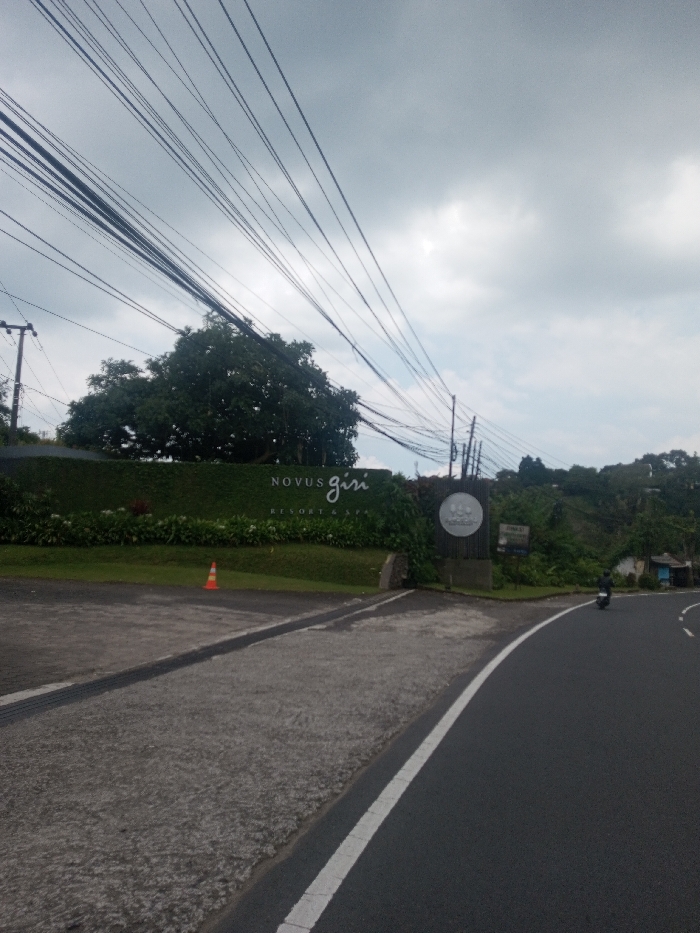 The image displays an exterior scene featuring a road curving past the entrance of "Novus Giri Resort & Spa" under an overcast sky. The location is Puncak, West Java, Indonesia, as indicated by the resort signage.
In the foreground, an asphalt road curves from the lower right towards the center, marked by a solid white line on its inner edge. To the left of the road is a concrete shoulder, containing a dark, rectangular drainage grate.
The midground on the left features the entrance to the resort. A prominent sign integrated into a tall, dense green hedge spells "NOVUS GIRI RESORT & SPA" in white lettering. To the right of this sign, a circular white sign with a logo is affixed to a stone pillar, partially obscured by foliage. An orange traffic cone rests on the concrete driveway leading into the resort. Above the hedge and signs, a large, mature tree with dark green foliage is visible. Numerous black utility cables traverse the upper portion of the image, running diagonally across the cloudy sky from left to right, supported by utility poles.
On the asphalt road in the midground, a single individual on a black scooter is observed moving away from the viewer, appearing on the right side of the road. The background reveals vegetated hills with scattered small structures under a grey, cloudy sky.