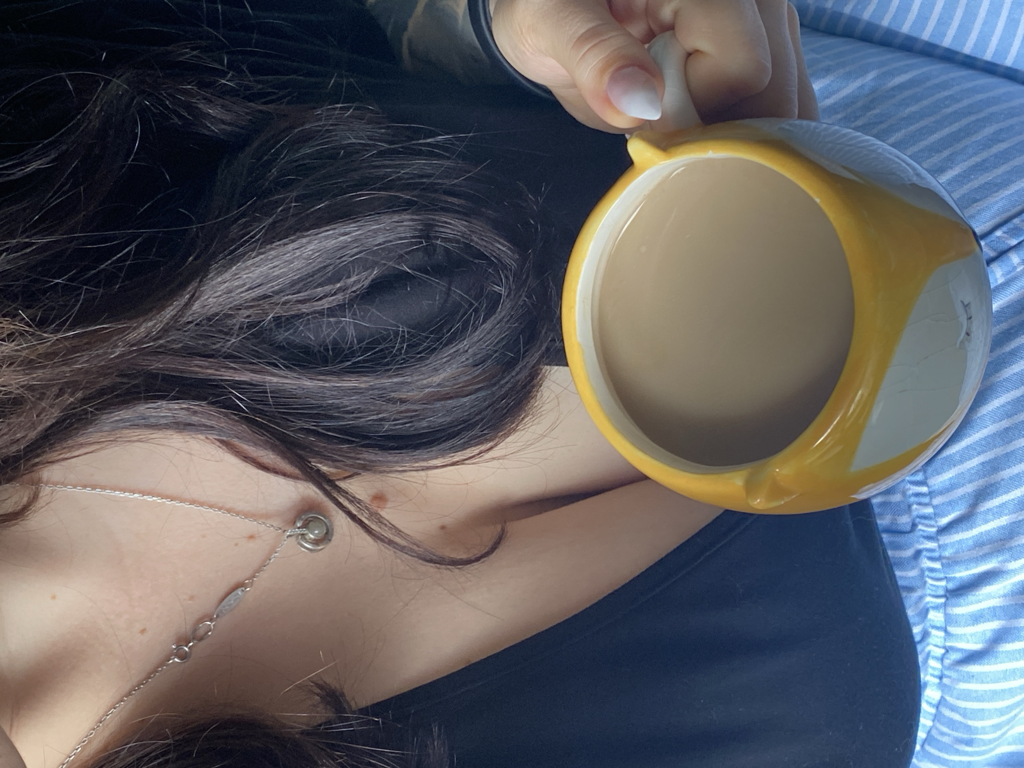 This close-up, high-angle photograph, taken in Chaikovsky, Russia, features a person holding a mug in a relaxed setting. The subject's dark, wavy hair dominates the left side of the frame, partially obscuring her face. She wears a dark, likely black, top. Around her neck is a delicate silver chain necklace with a small, round charm. Her fair skin shows a small mole near her collarbone.
Her hand, with manicured nails displaying a white ombre or French tip style, holds a yellow and white mug. The mug contains a light brown liquid, consistent with coffee or tea with milk. The mug itself has an irregular, possibly textured or painted, yellow and white design. To the right of the person, a light blue fabric with thin white stripes is visible, suggesting a patterned pillow, blanket, or another piece of clothing. The overall scene conveys a calm, domestic moment, possibly during a leisure activity like drinking a beverage. There is no visible text in the image. The lighting is soft and ambient, characteristic of an indoor environment.