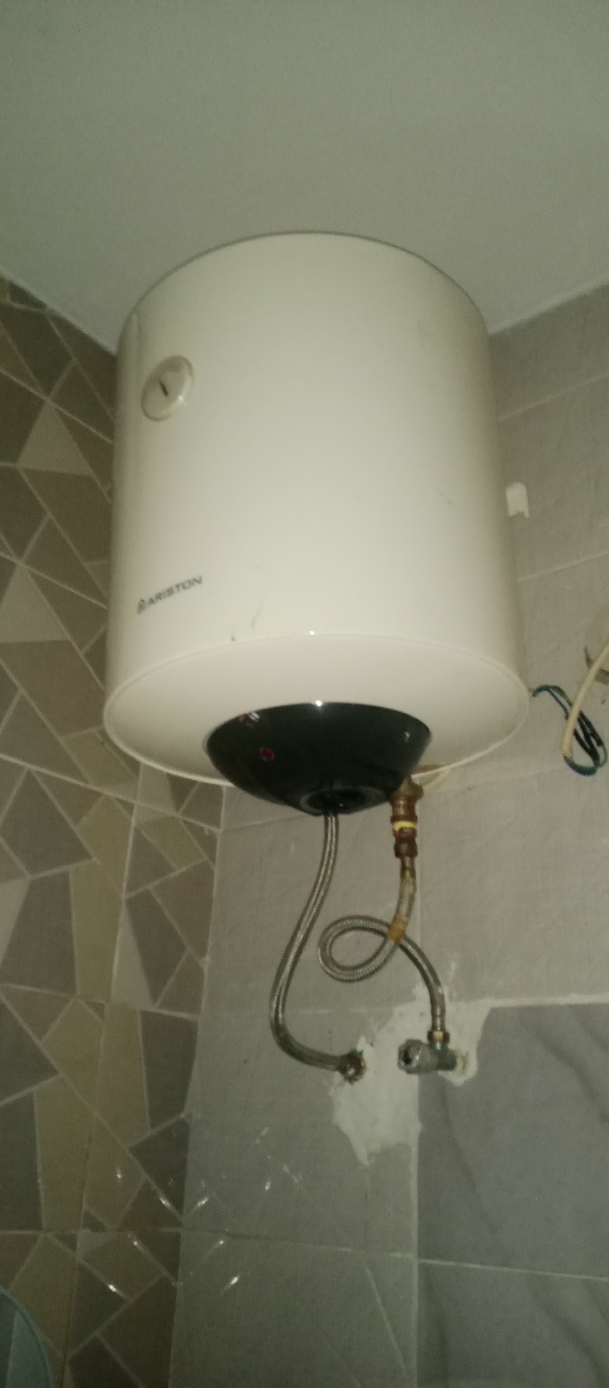The image shows a white Ariston brand water heater mounted on a tiled wall, likely in a bathroom or utility area in Gwarinpa, Nigeria. The water heater is a cylindrical appliance with a black base. Two flexible metal hoses are connected to the bottom of the unit, one leading downwards and the other curving to the right, where it connects to a faucet. The walls are covered in geometric patterned tiles in shades of beige and gray. An electrical wire is visible connected to the side of the water heater, with blue and green wires. The ceiling is visible at the top of the image. The overall environment appears to be a residential or commercial building interior.