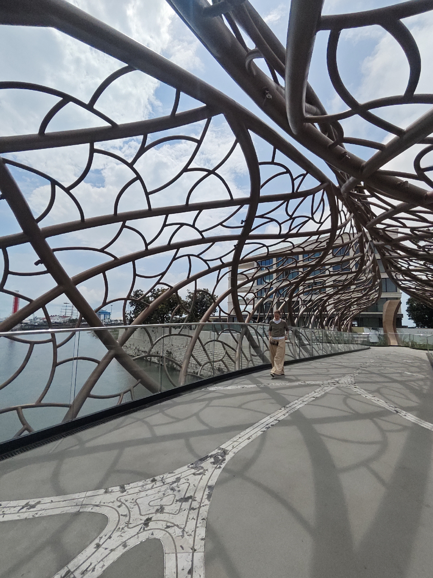 A woman is walking across the Figurnoye Kataniye Bridge in Sochi, Russia. The bridge's unique architectural design, featuring interwoven metal elements resembling branches, creates a striking visual effect. The sky is partially cloudy. The woman is wearing a light-colored outfit. A body of water is visible on the left, and buildings are in the background. The bridge surface has an intricate design.