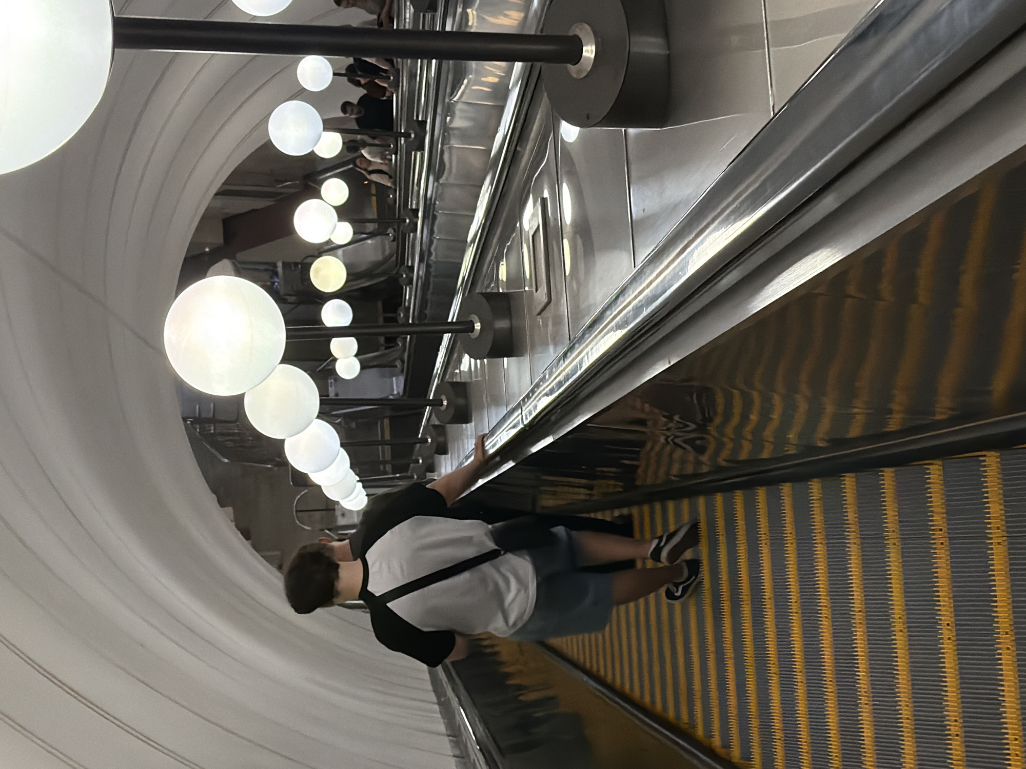 A person is ascending a wide escalator in a Moscow Metro station. The station features a distinctive arched ceiling and is illuminated by a series of spherical, globe-like light fixtures mounted on dark poles. The escalator steps are gray with yellow warning stripes. The person, seen from behind, is wearing a white t-shirt with black sleeves, blue shorts, and black sneakers with white laces. A black crossbody bag is slung over their shoulder. Another person is partially visible behind them. Other people are seen on adjacent escalators in the background, also ascending. The overall impression is of a modern, well-lit, and functional underground transit environment.