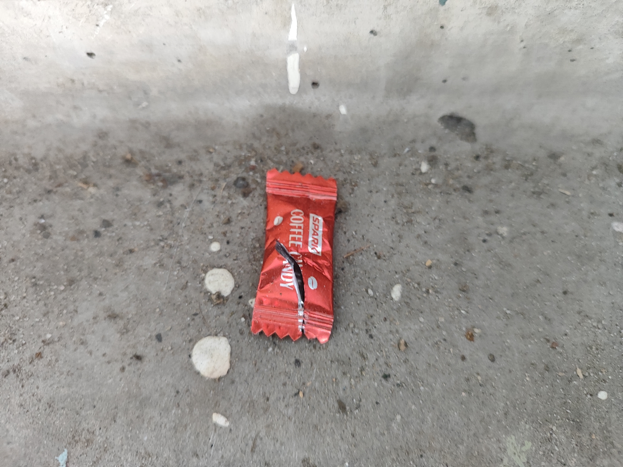 On a gray concrete surface in Unknown, Unknown, lies a discarded red candy wrapper with the words "Spark Coffee Candy" printed on it. The wrapper is torn open, indicating the candy has been consumed. The surrounding concrete surface is uneven and has various blemishes, including white splatters and dark spots.