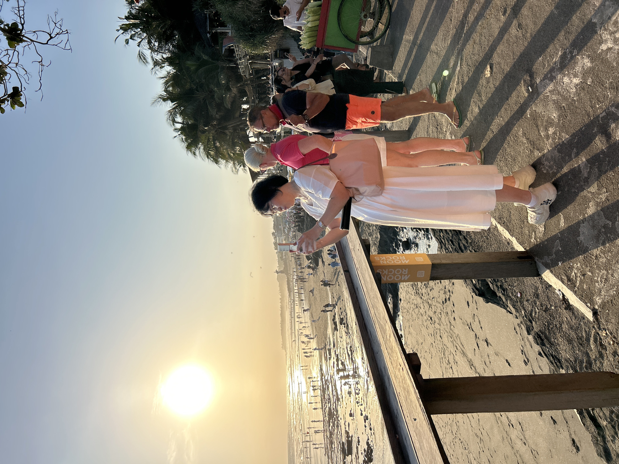 The image captures a sunset scene on a beach in North Kuta, Indonesia. In the foreground, a woman in a white dress is taking a photo with her phone. To her right, two other people stand, with a man wearing orange shorts and a woman in a pink shirt. Further back, other individuals are visible, some looking at their phones. A vendor's cart laden with corn is parked on the right side of the frame, and a group of people are gathered around it. The beach itself is populated with many individuals enjoying the late afternoon. Palm trees and a building with a thatched roof are visible in the background. The sun is low in the sky, casting a warm glow and long shadows across the scene.