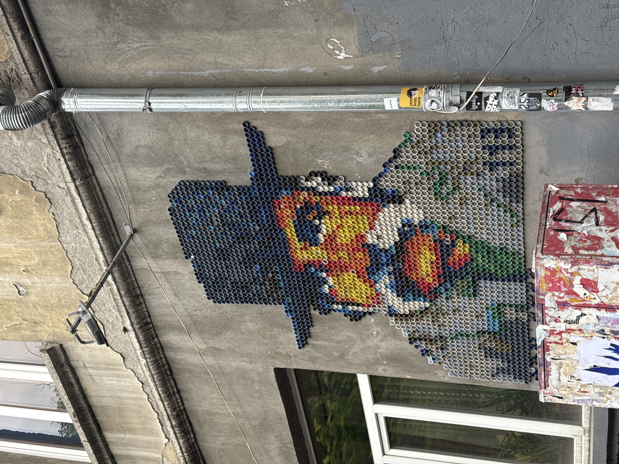 The image captures a street art mosaic affixed to a building exterior in Yerevan, Armenia. The mosaic, constructed from small, cylindrical objects (possibly bottle caps or tubes), depicts an abstract colorful design, featuring blue, red, yellow, green, and white elements. Above the mosaic runs a horizontal metal pipe adorned with an array of stickers. Adjacent to the mosaic, on the right, is a weather-beaten metal structure, possibly a mailbox or electrical box, covered in layers of peeling paint and remnants of posters or advertisements. The building itself exhibits a weathered concrete facade. Windows with white frames are visible below the artwork, reflecting greenery from outside. The overall scene presents an urban setting with evidence of age, wear, and artistic expression.