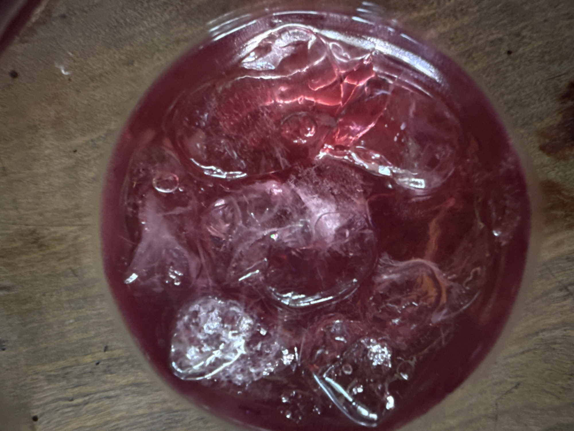 A top-down, slightly blurred shot shows a clear glass filled with a reddish-purple liquid and numerous ice cubes. The glass sits on a wooden surface with visible grain. The ice cubes are mostly clear, with some showing reflections and refractions of the colored liquid. No city or specific location is discernible. No people, actions, or interactions are depicted. The environment consists solely of the wooden surface and the glass with its contents.
