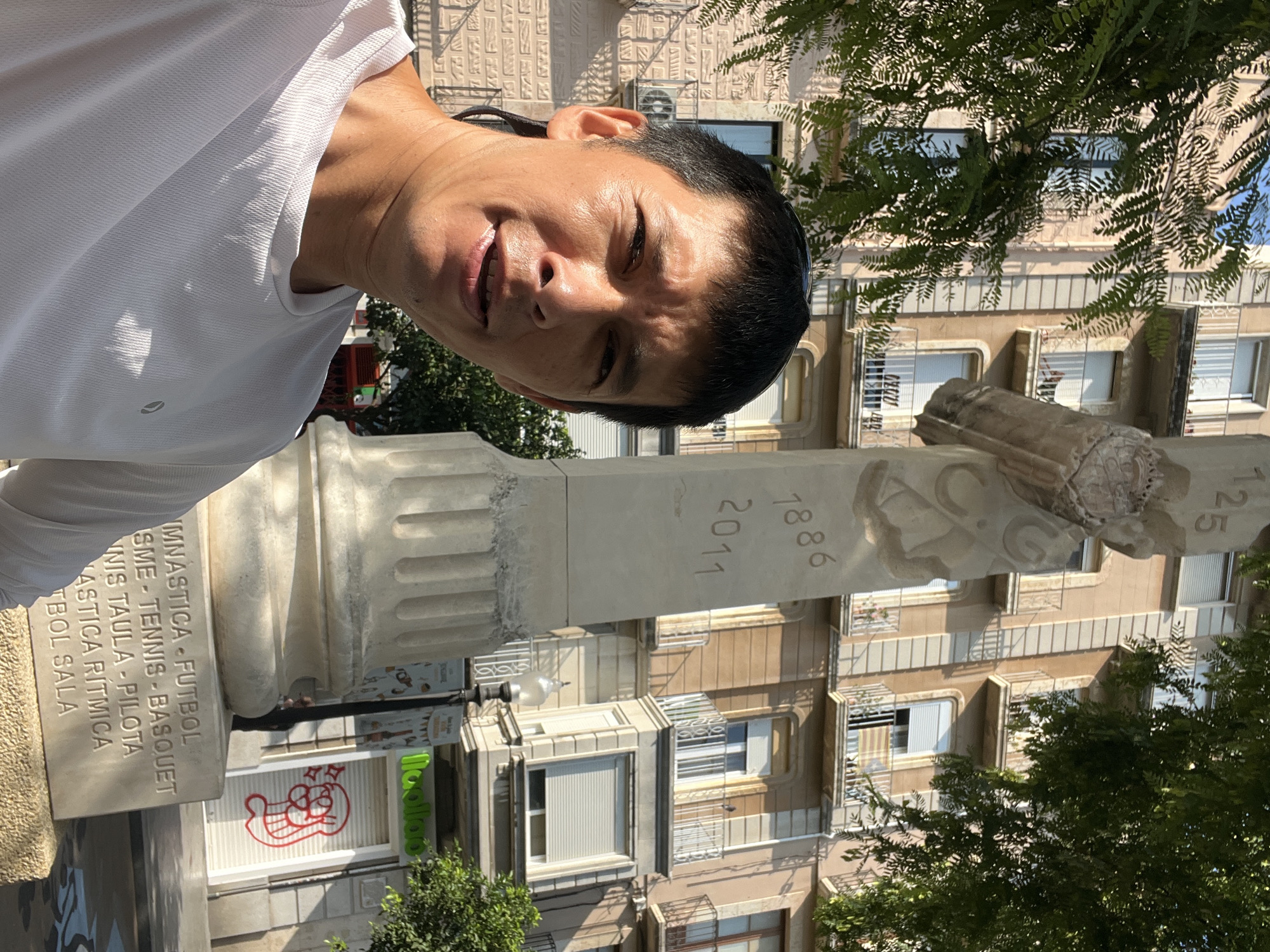In Tarragona, Spain, a man wearing a white t-shirt is taking a selfie outdoors. He is positioned to the left of a large, light-colored monument with carved inscriptions and decorative elements. Behind the monument and the man, a multi-story building with beige and brown facades stands, featuring multiple balconies with white railings. The building has several windows, some of which are covered by blinds, and one balcony displays a Catalan flag. Lush green trees frame the scene from the top left and right, casting dappled shadows. At the base of the monument, a stone plinth displays text in capital letters, listing various sports. To the right of the monument, a street lamp is visible, and behind it, the facade of a closed shop is adorned with graffiti. The sky is clear and blue, suggesting a sunny day.