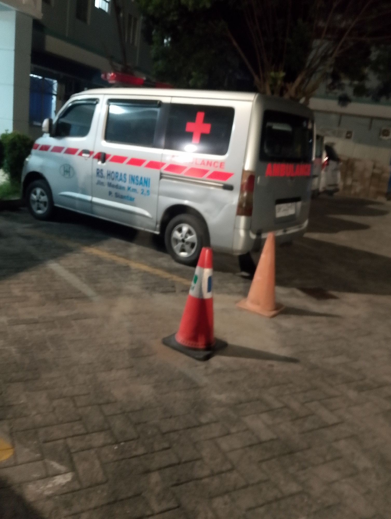 The image captures a white ambulance parked on a paved surface, possibly a parking lot, in front of a building at night. The ambulance has red stripes along its side and a red cross symbol. Text on the ambulance indicates "RS. HORAS INSANI" and "AMBULANCE". Two traffic cones are visible in the foreground, one orange and one red and white. A tree is present behind the ambulance. The location is identified as the City of Pematang Siantar, Indonesia, based on the text on the ambulance, "RS. HORAS INSANI" which is a hospital in Pematang Siantar, and the address "Jl. Medan Km. 2.5 P. Siantar." This suggests the scene is at or near RS. Horas Insani in Pematang Siantar.