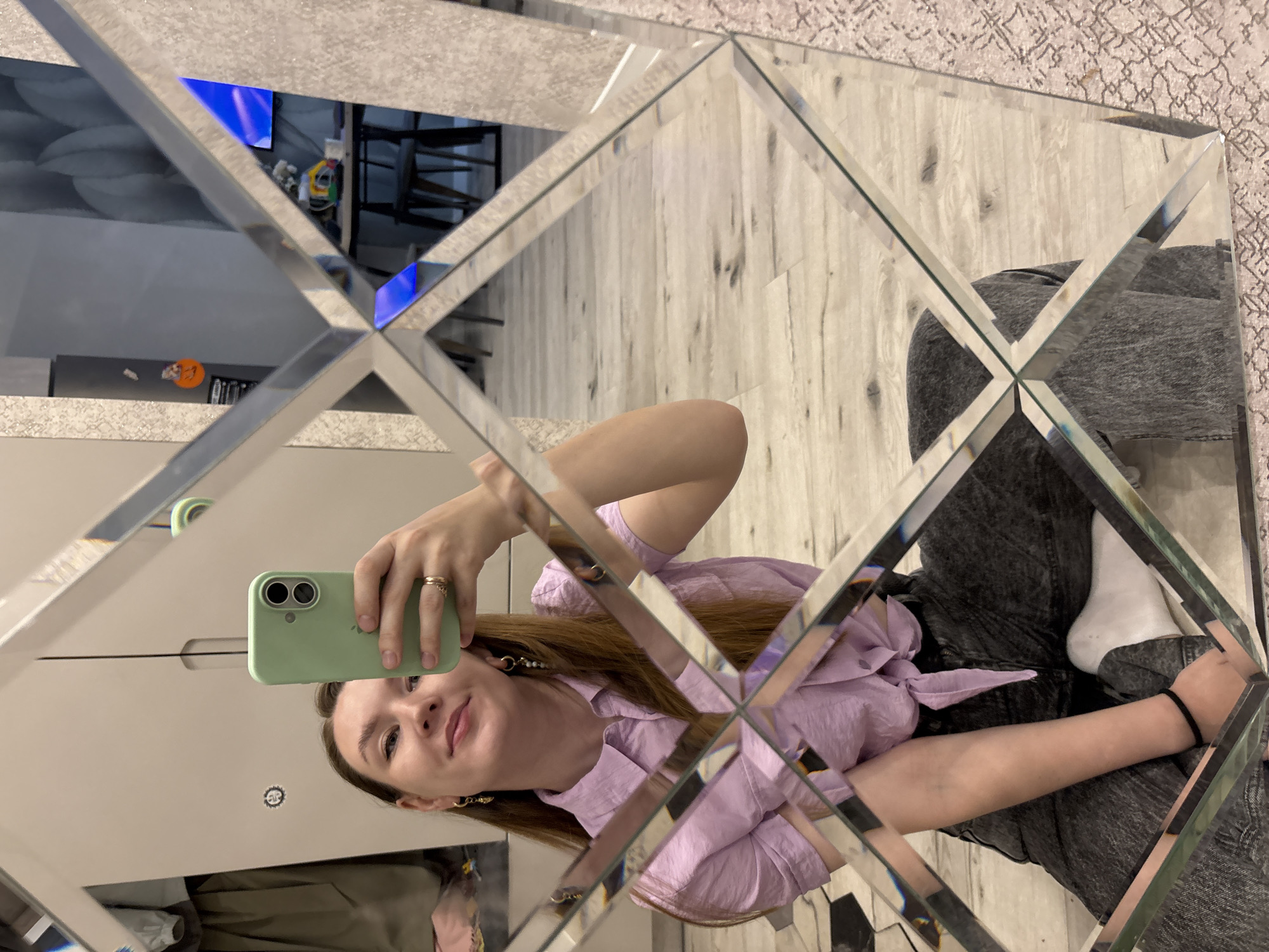 A young woman is captured in a mirror selfie, reflecting her image in a decorative, multi-faceted mirror. The mirror is composed of diamond-shaped segments with beveled edges, fragmenting and distorting the reflections of the room around her. She has long, reddish-brown hair and is looking directly into the camera with a gentle, faint smile. She wears a light purple, textured blouse with voluminous sleeves, dark wash jeans, and a white sock on her visible right foot. Her right hand, adorned with a ring, holds a light green iPhone with a dual camera system, which she uses to take the photo. She is also wearing prominent hoop earrings.
The reflections in the mirror offer glimpses of an indoor setting in Izhevsk, Russia. A light-colored wooden floor is visible in several segments, suggesting a modern interior. Other segments show parts of a room with furniture, including what appears to be chairs and possibly a table, and a wall or surface with a subtle geometric pattern in the background. A distinctive blue light, possibly from a television screen or an LED fixture, is also visible in some of the mirror segments. The overall impression is of a casual moment captured indoors, likely during the day or evening, given the ambient lighting. No discernible text is visible in the image.