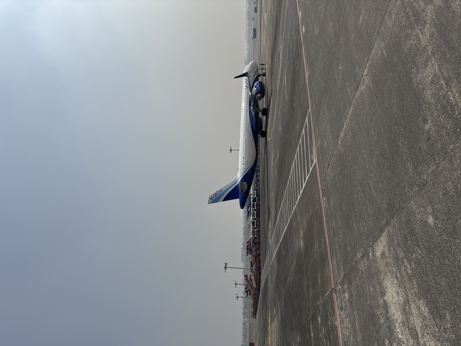 An Indigo airplane sits stationary on an airport tarmac. The plane is painted with a blue and white color scheme, and the Indigo logo is visible on the tail. The tarmac is gray and marked with white and red lines. Background shows a row of airport vehicles and equipment. The sky is overcast and pale.