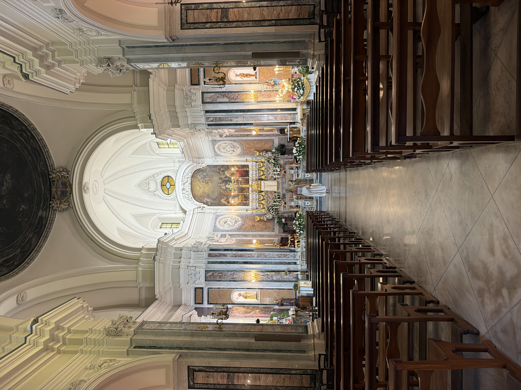 The image shows the interior of a church in Ischia, Italy. The view is from the back of the church, looking toward the altar. Rows of wooden pews line both sides of the central aisle. The interior of the church is ornately decorated with marble columns, paintings, and statues. Natural light streams in through windows in the walls. There is a person in the aisle, near the altar. The church is empty except for this individual.