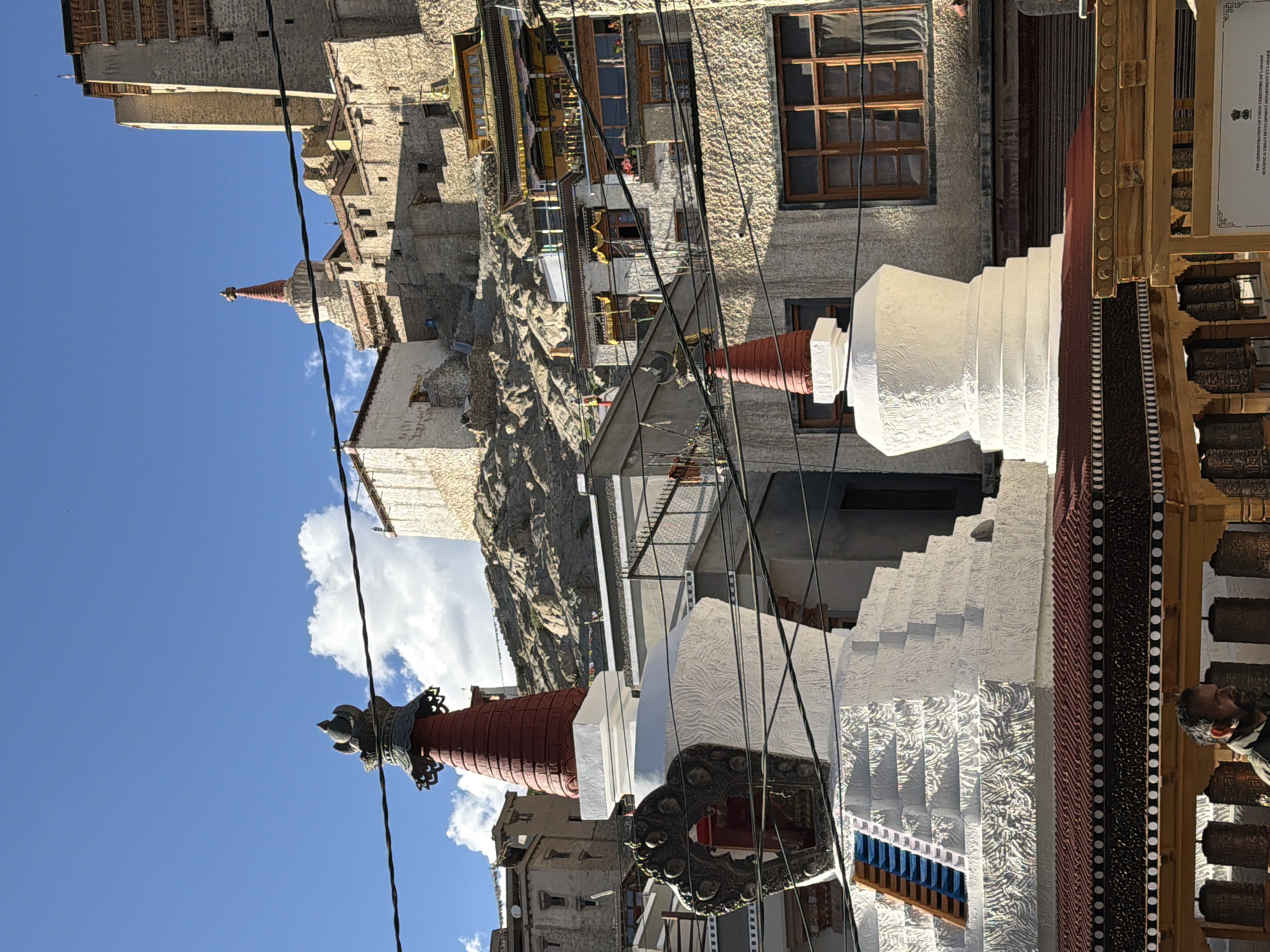 This is a daytime, eye-level, medium shot of the Leh Palace complex in Leh, India, a historic structure built in the 17th century. The scene is dominated by a clear blue sky with scattered white clouds. In the foreground, a large white stupa with intricate carvings stands prominently, topped with a dark, ornate finial. To its right, a smaller white stupa is visible. Behind the stupas, the stone buildings of the Leh Palace rise up the hillside, characterized by their rough-hewn stone walls, wooden window frames, and layered architecture. Various Tibetan prayer flags are strung across the scene. A man is partially visible at the bottom of the frame, looking to the left, standing near prayer wheels. Wires are strung across the upper portion of the image, obscuring parts of the sky and the distant buildings.