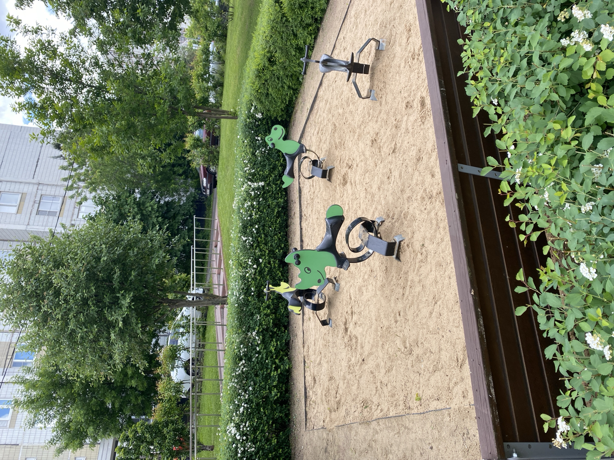 The image shows a children's playground in Khimki, Russia. The playground surface is sand, and there are several play structures, including three green and black dinosaur spring riders and a single spring rider with three seats. A low wooden fence forms one side of the playground, with bushes growing alongside it. Trees and buildings are visible in the background, indicating an urban environment.