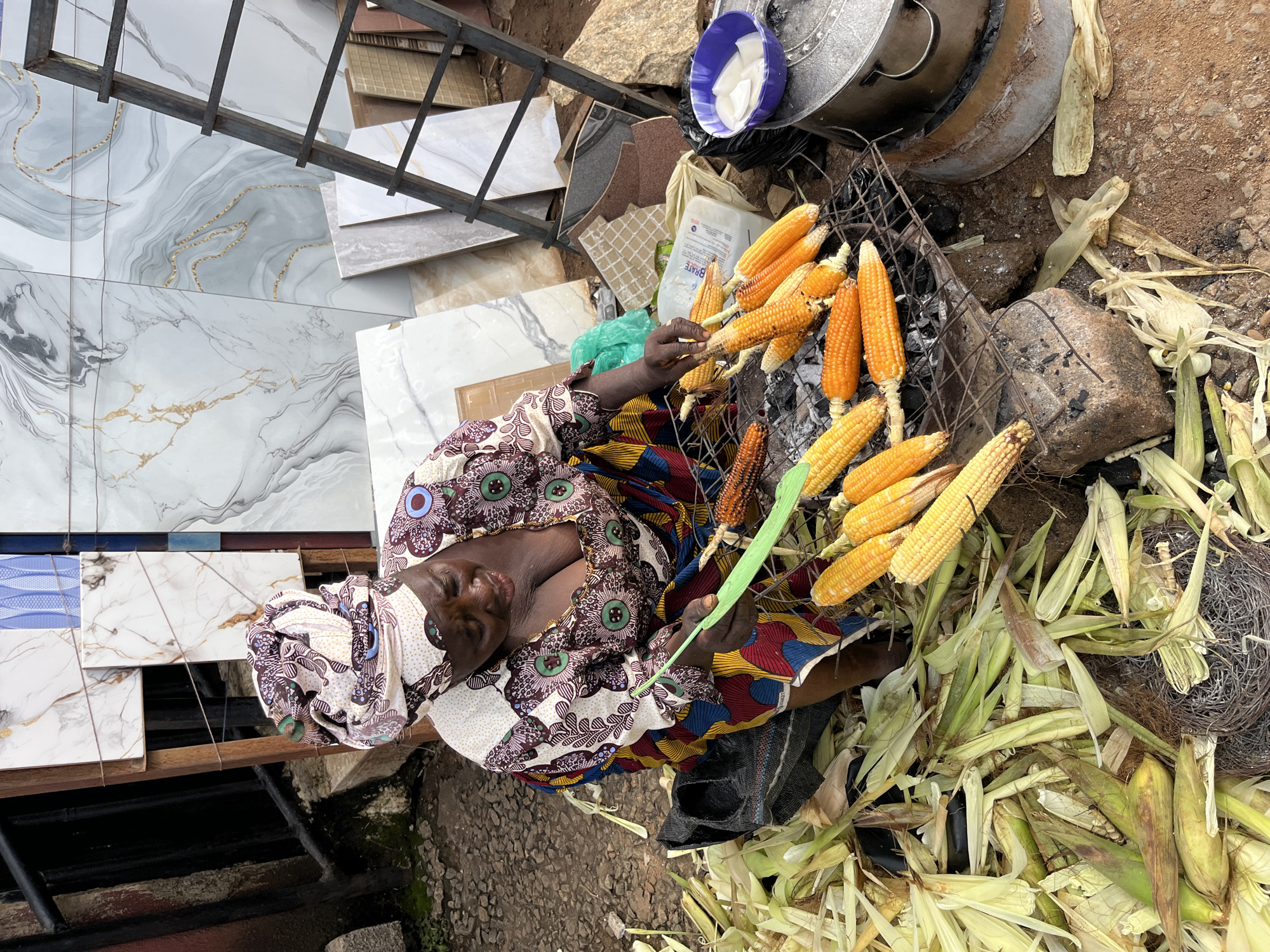 An overhead, close-up, eye-level shot shows a Black woman in traditional clothing and headwrap at an outdoor market stall. She is dressed in a vibrant, patterned wrap dress and headscarf. The woman is tending to a charcoal grill laden with corn on the cob, some of which are golden yellow and others slightly more toasted. She holds a green utensil, possibly for turning the corn. Around her, there are piles of husked corn, corn husks, and what appear to be tiles or slabs of stone for sale. A large metal pot and a blue bowl containing white slices, possibly coconut, are also visible. The scene suggests a food stall at a Nigerian market, likely during daytime, with the ground appearing to be dusty earth. The woman's eyes are closed and her head is tilted back, giving an impression of a moment of rest or quiet contemplation amidst her work. The overall atmosphere is one of vibrant commerce and daily life.