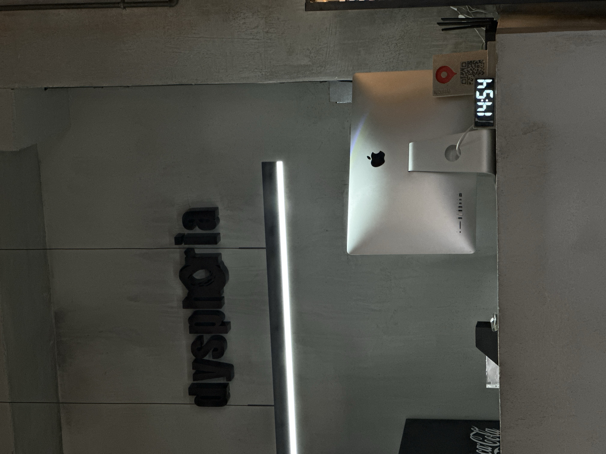 The image shows a vertical view of a section of an interior wall with the name "Dyshoria" in vertical black letters. Beside the letters, a vertical LED light fixture is present. An Apple iMac computer is mounted on the wall, and a digital clock displaying "14:54" is adhered to the side of the computer. The background wall is concrete. A portion of a "Coca-Cola" logo is visible in the lower right. While the precise location cannot be definitively determined from this limited visual information, the presence of the Dyshoria logo would indicate that this picture was taken at the Dyshoria store in Moscow, Russia.