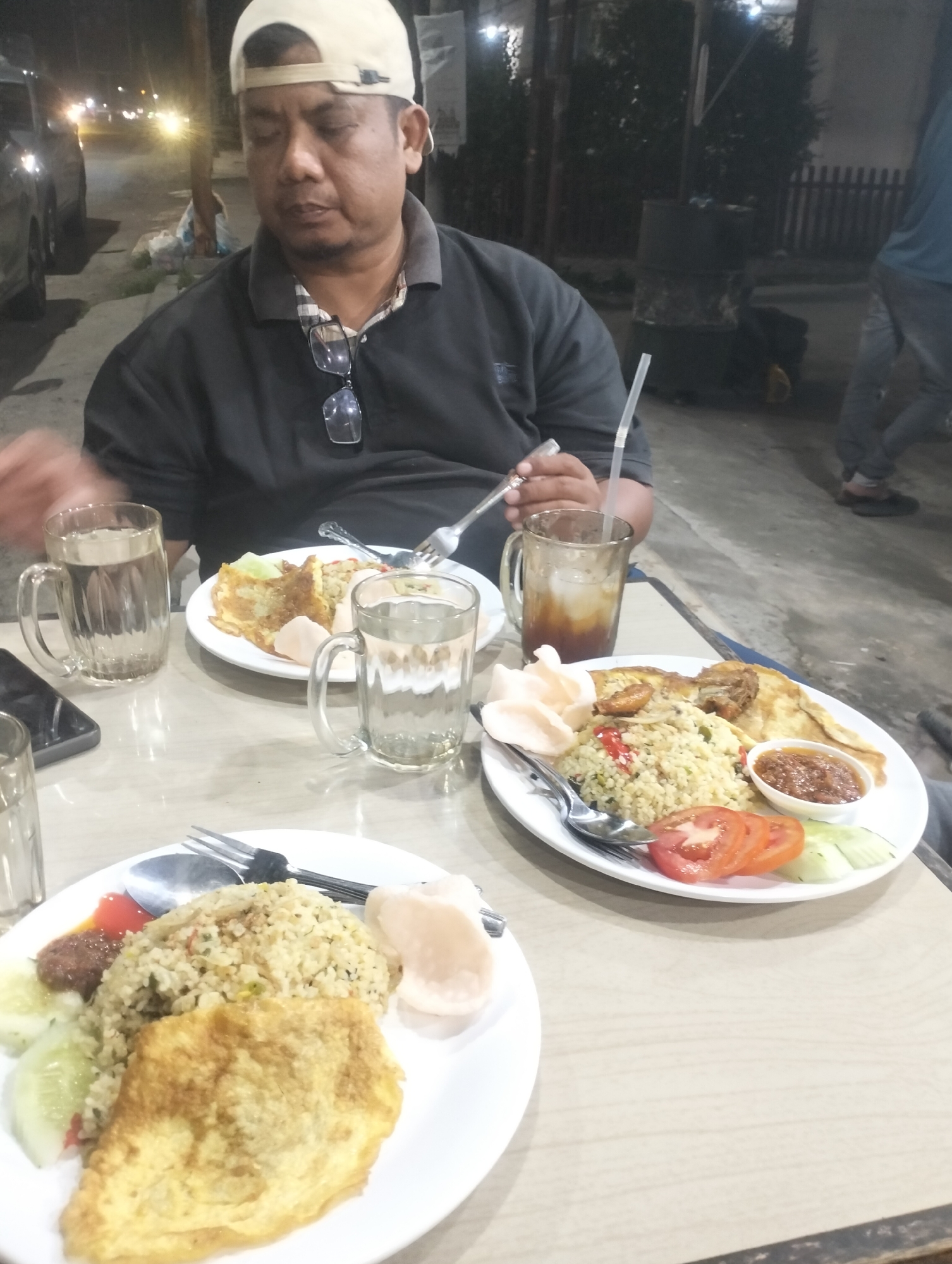 A man wearing a light-colored baseball cap and a dark polo shirt sits at a table with two plates of food and two glasses of water. He has glasses hanging from his shirt. One plate contains fried rice, an omelet, sliced tomatoes, and cucumbers. The other plate has a similar arrangement of food. A glass of iced tea with a straw is also on the table. The scene appears to be an outdoor dining area at night, possibly a restaurant or street food stall in Lhokseumawe, Indonesia, indicated by the streetlights and the style of the food. The man is looking down at the food on his plate.